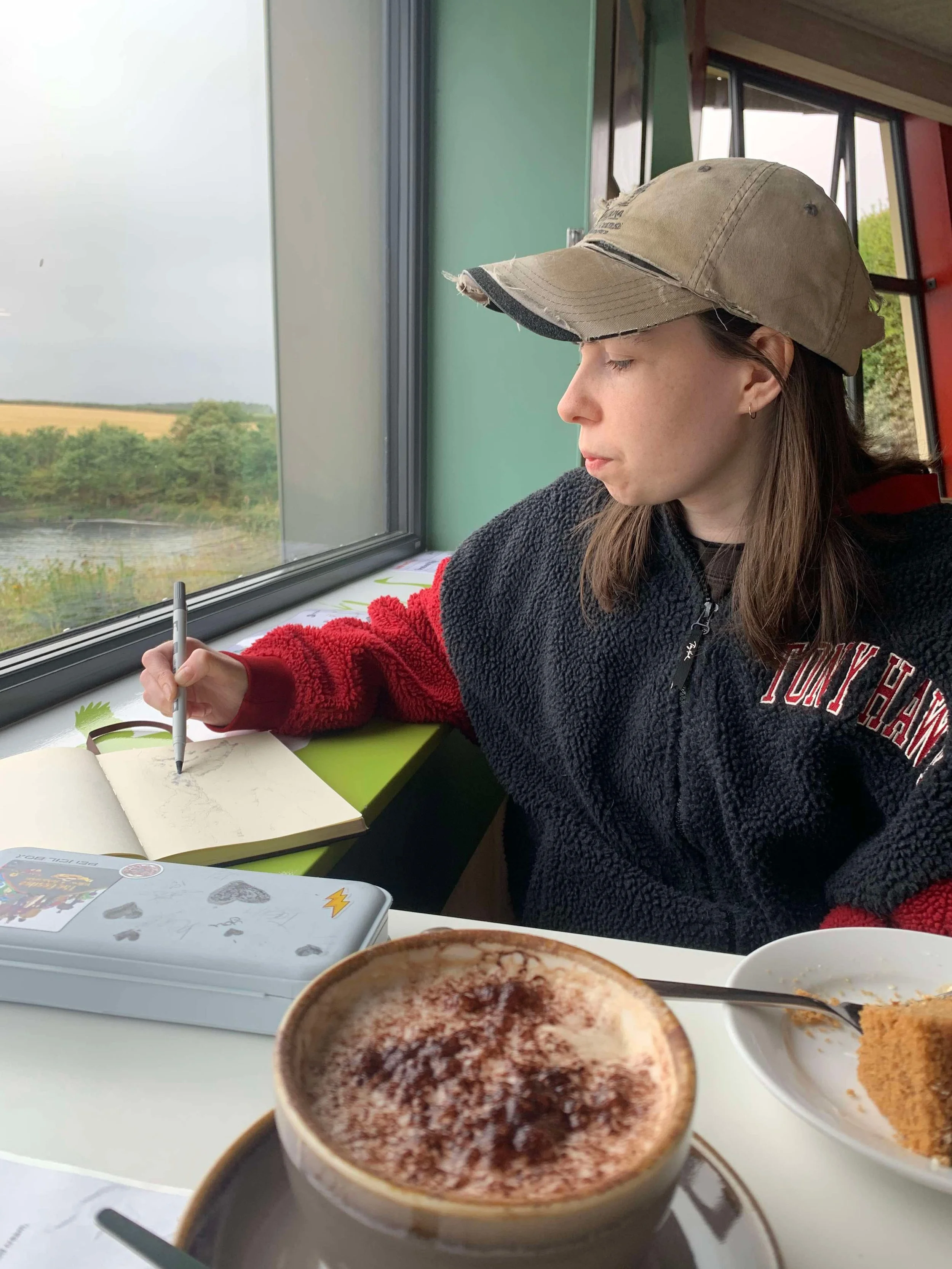 A woman with brown hair, wearing a beige cap and a black and red fleece jacket, sitting by a window and drawing in a sketchbook with a pen. On the table in front of her are a cup of coffee with foam and chocolate, a slice of cake, and a white plate. Outside the window, there is a view of a field and trees.