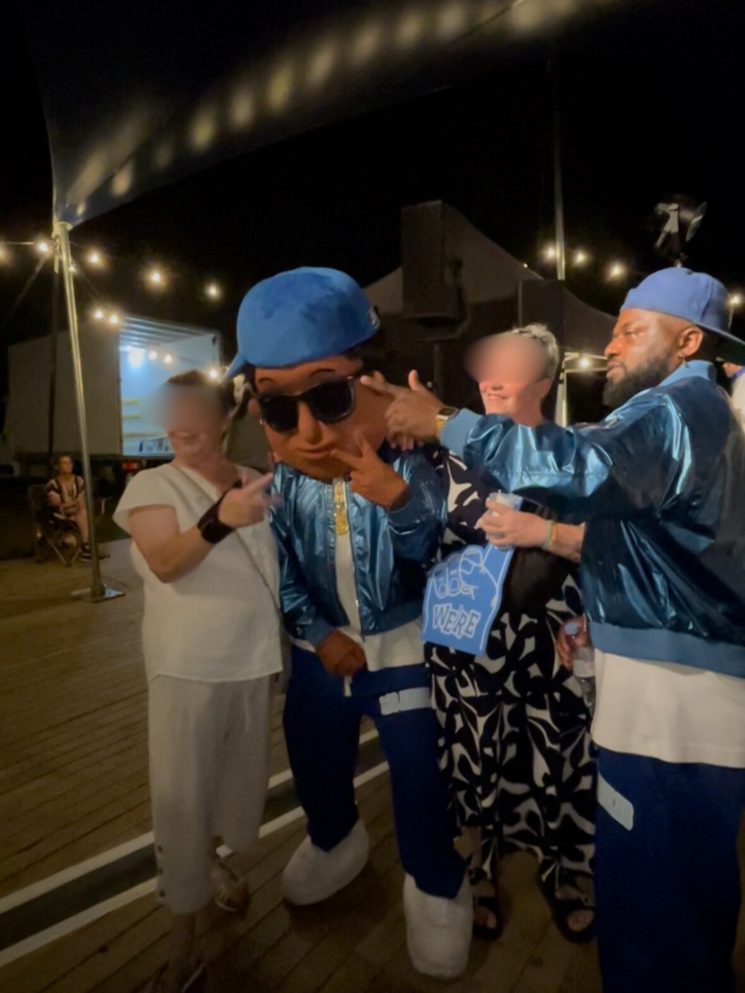 Groupe de personnes déguisées en figures célèbres ou caricatures, dans une ambiance nocturne avec des lumières, échangeant des gestes dans une ambiance festive. Concert de El Loco avec Scotti la mascotte à Cesson-Sévigné.