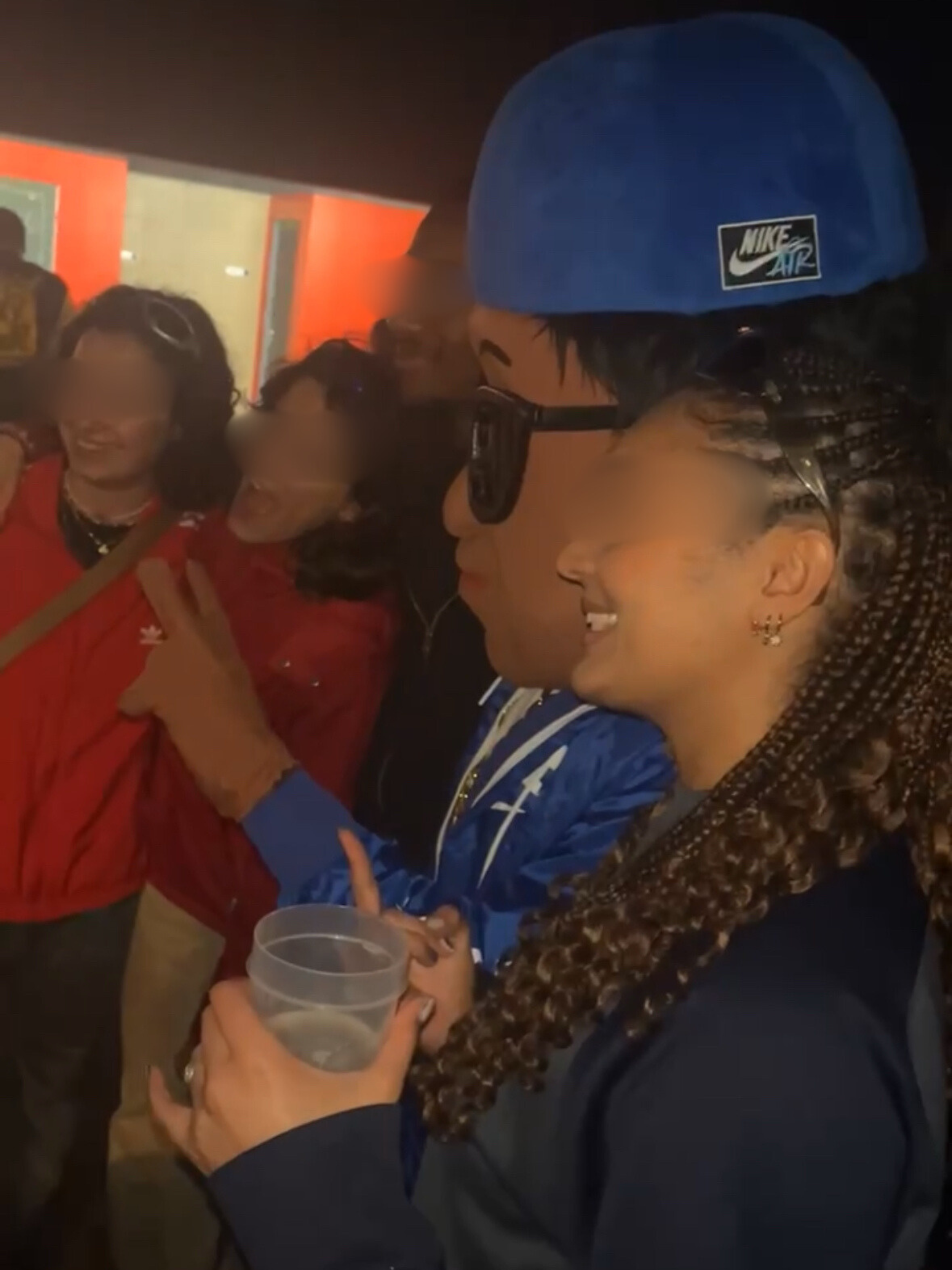 Groupe de personnes souriantes en intérieur, la mascotte scotti portant une casquette Nike Air, des lunettes de soleil et tenant un verre d'eau, entourée d'amis posant pour la photo.