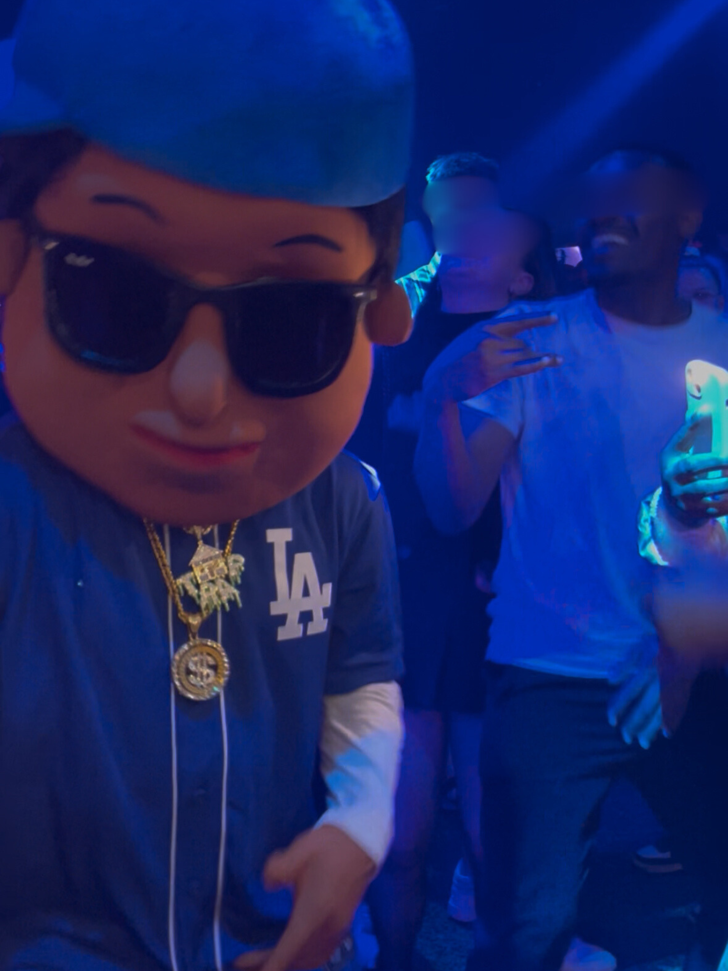 Personnage en costume avec une grosse tête portant des lunettes de soleil, portant des abréviations de Los Angeles sur sa veste, dans un environnement sombre avec un éclairage bleu. C'est Scotti la mascotte en discothèque.