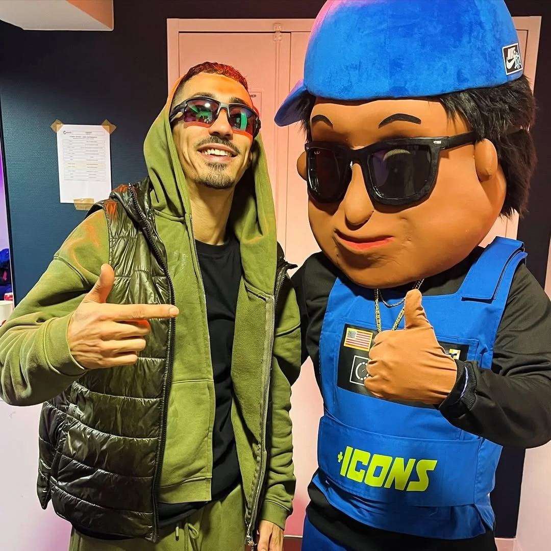 Un homme souriant portant des lunettes de soleil et un hoodie vert, pointant vers un personnage mascotte habillé en tenue de police ou de sécurité. C'est le rappeur Stony Stone et la mascotte Scotti.