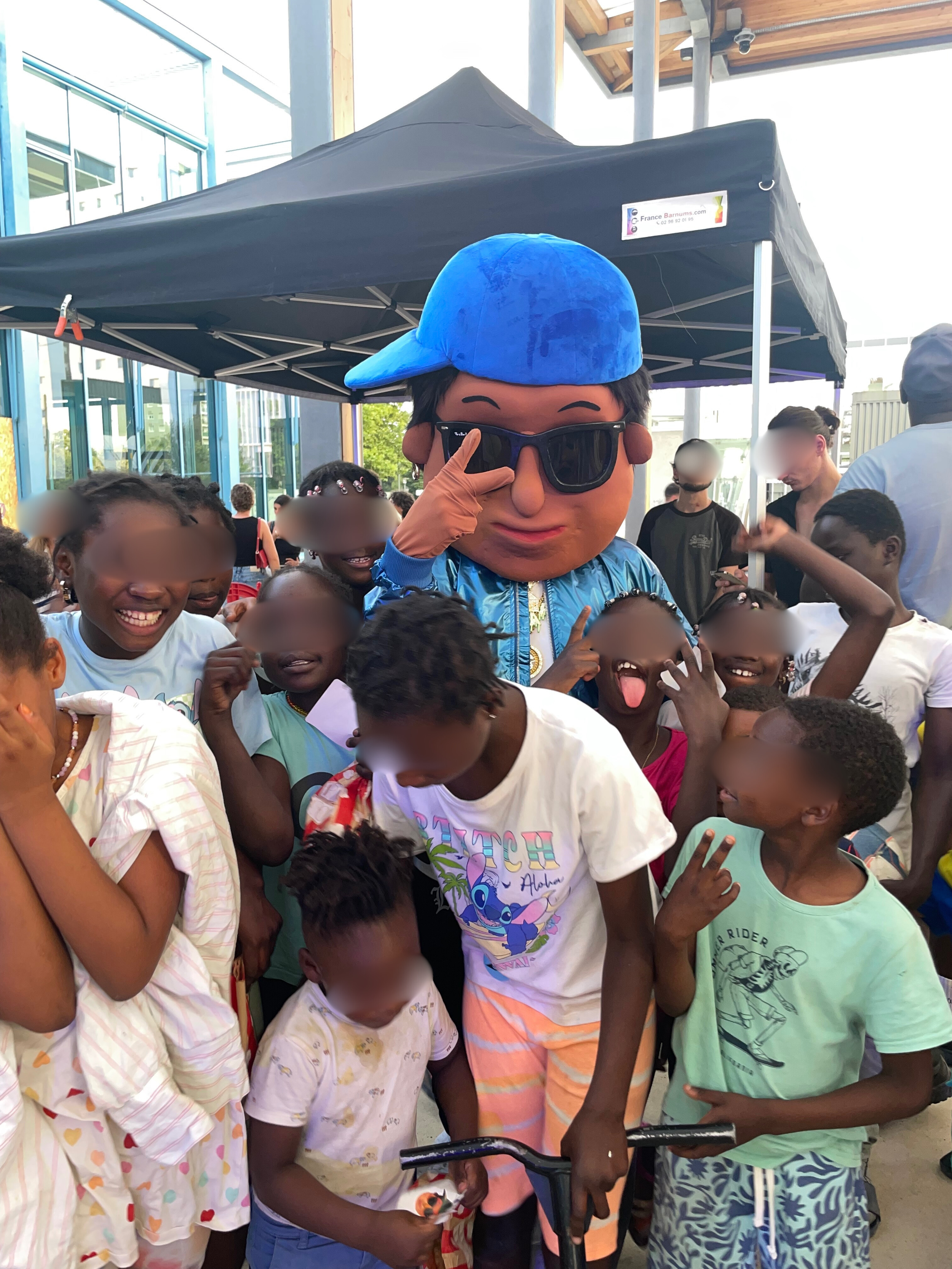 Un groupe d'enfants souriants et joyeux autour d'une mascotte géante en costume, sous une tente noire, lors d'un événement extérieur. C'est la mascotte Scotti dans le quartier du Blosne à Rennes