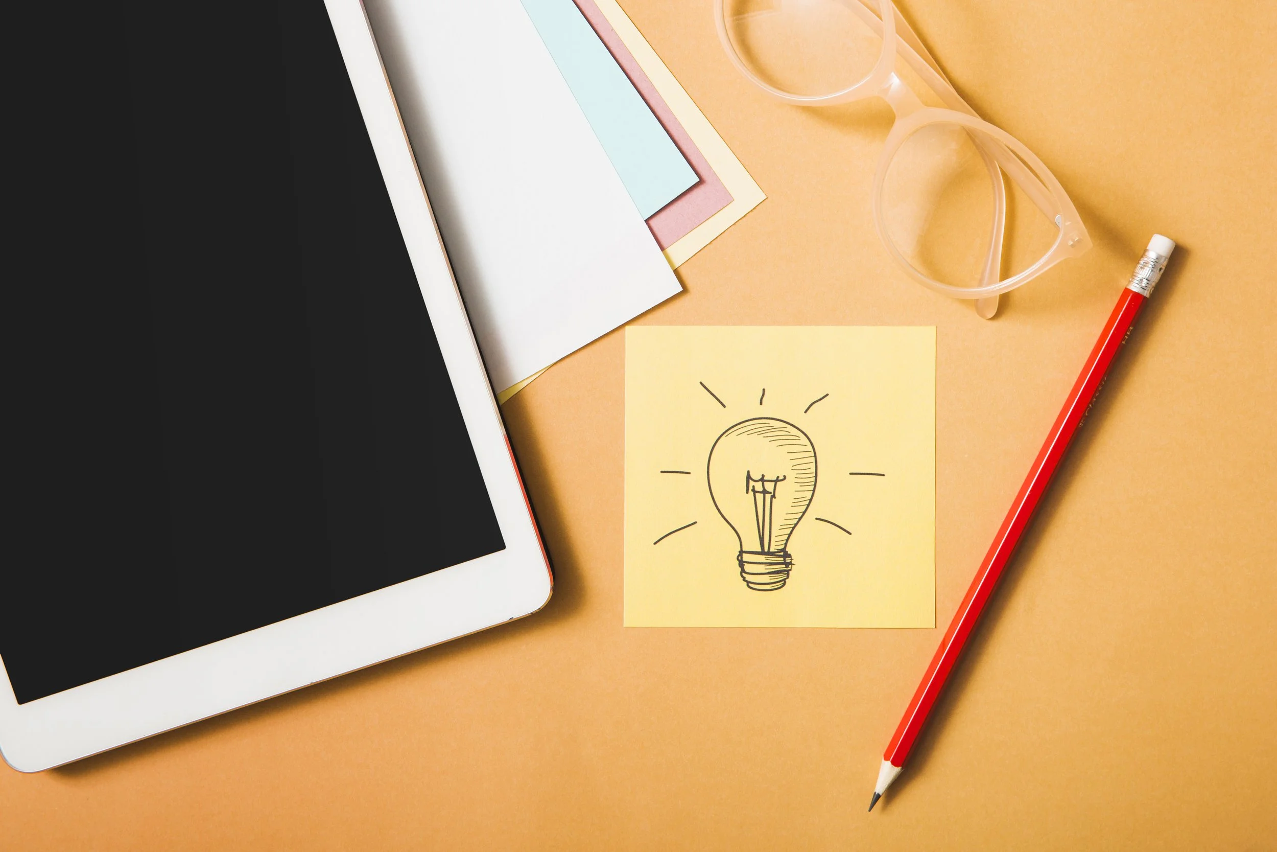 Une tablette, des papiers colorés, une paire de lunettes en plastique transparent, un crayon rouge, et un post-it jaune avec un dessin d'ampoule électrique sur un fond orange illustrant une idée.