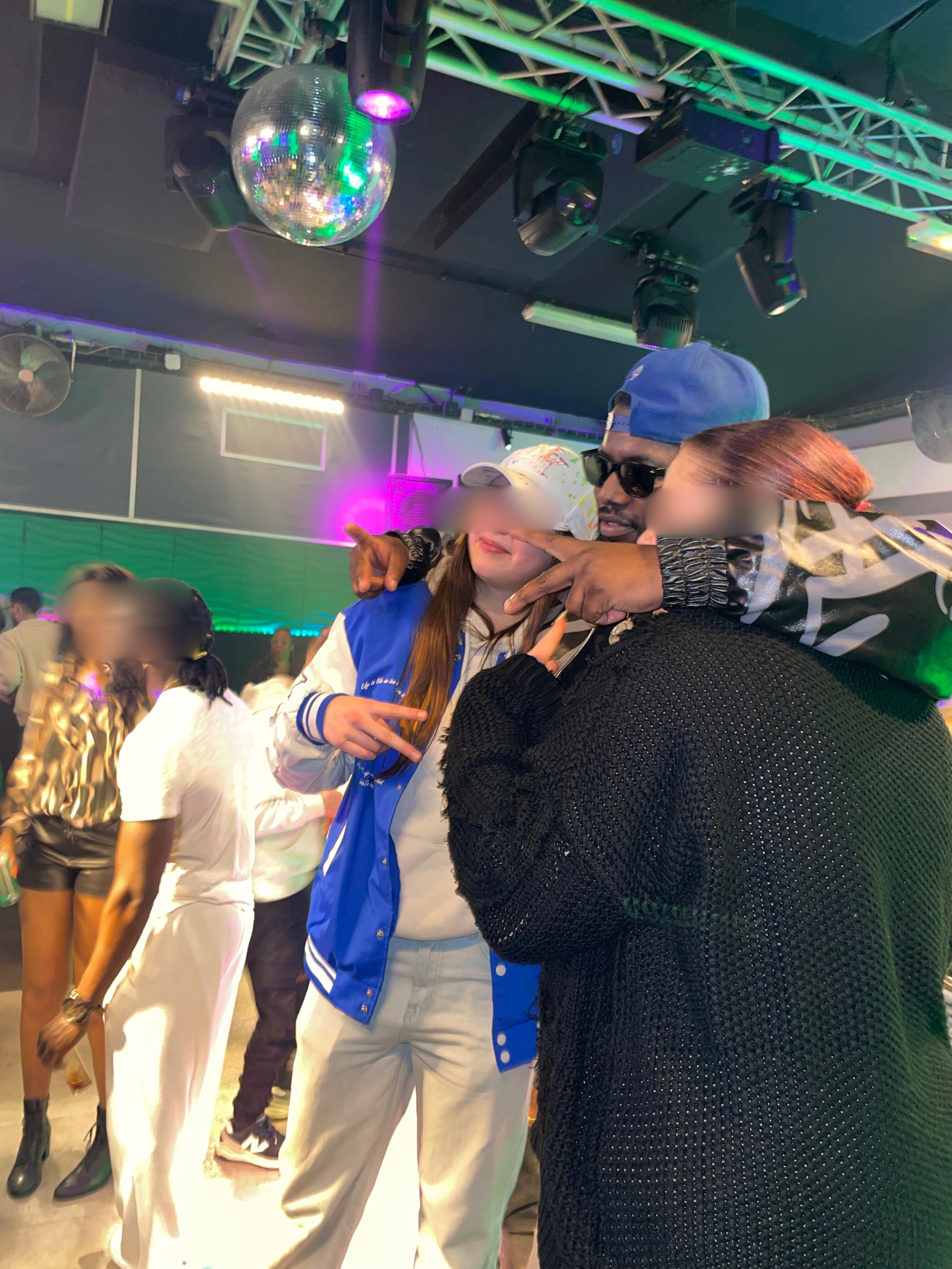 Trois personnes posent ensemble dans une discothèque, deux femmes et un homme. La scène est illuminée de lumières colorées, avec une boule disco au plafond. Le rappeur El Loco en concert dans la discothèque le Klub à Fougères.