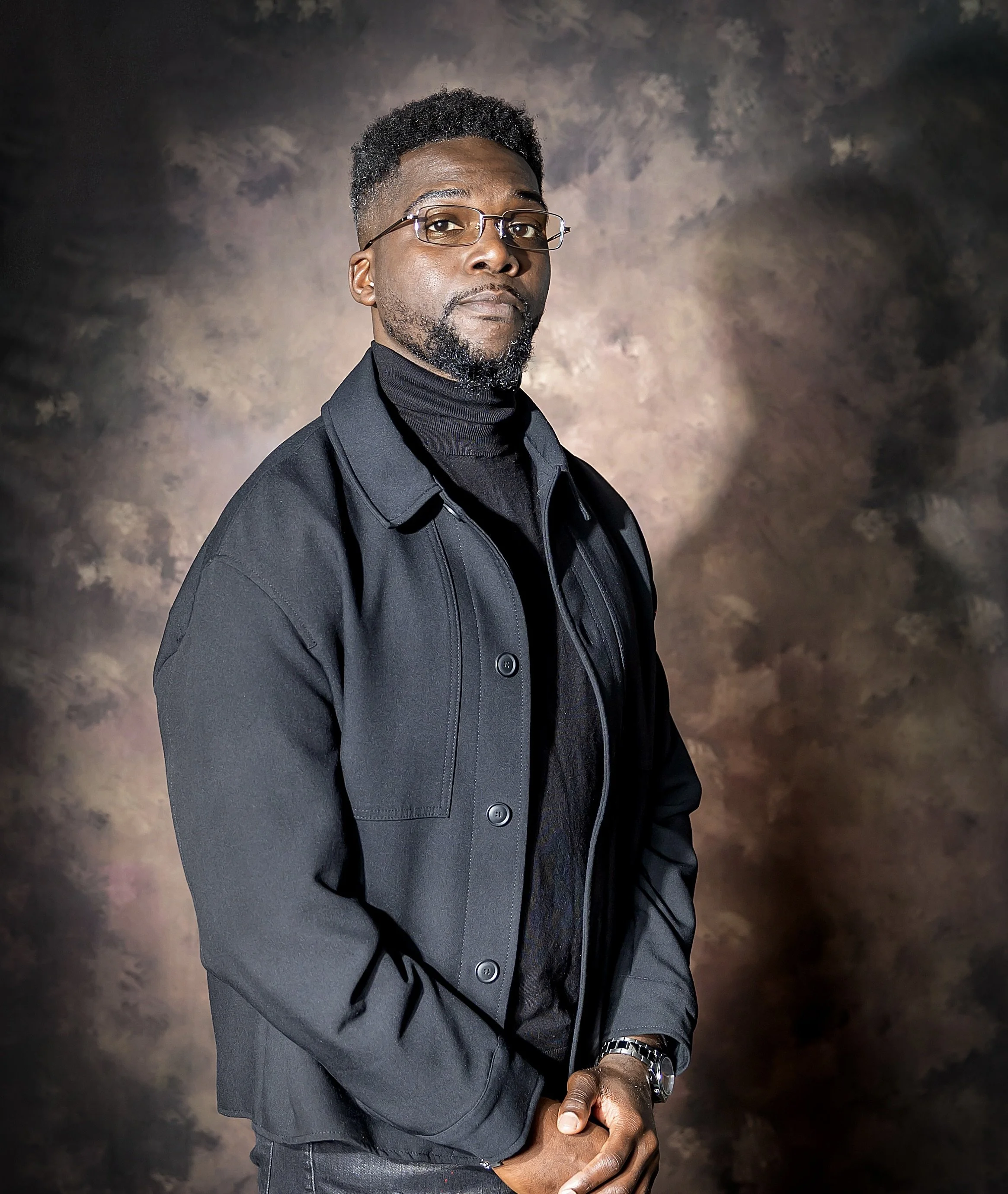 Gérant NLIK Marketing portant des lunettes, un pull noir et une veste noire se tient devant un fond brumeux. Il a la peau foncée et des cheveux courts et bouclés.
