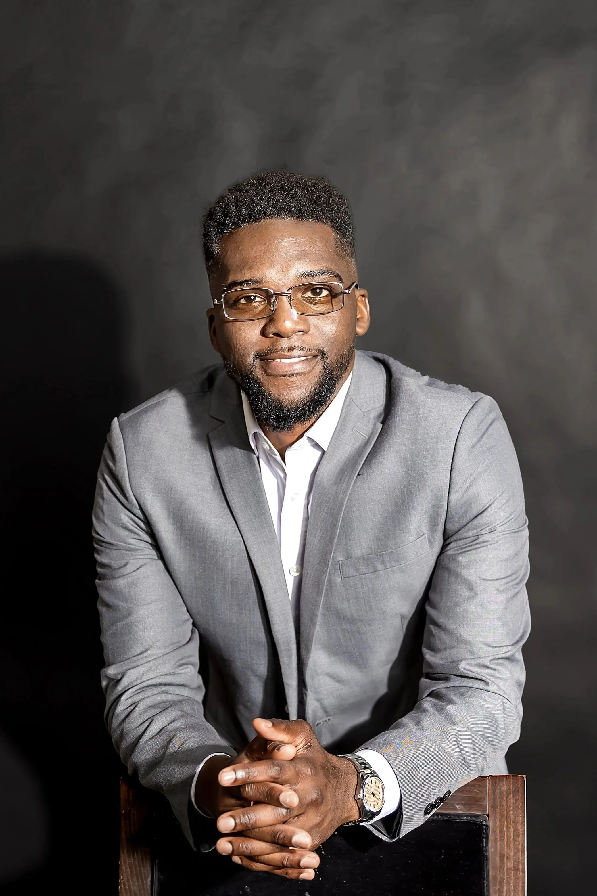 Une portrait d'homme d'affaires africain avec lunettes, portant un costume gris, assis avec les mains croisées, fond sombre.