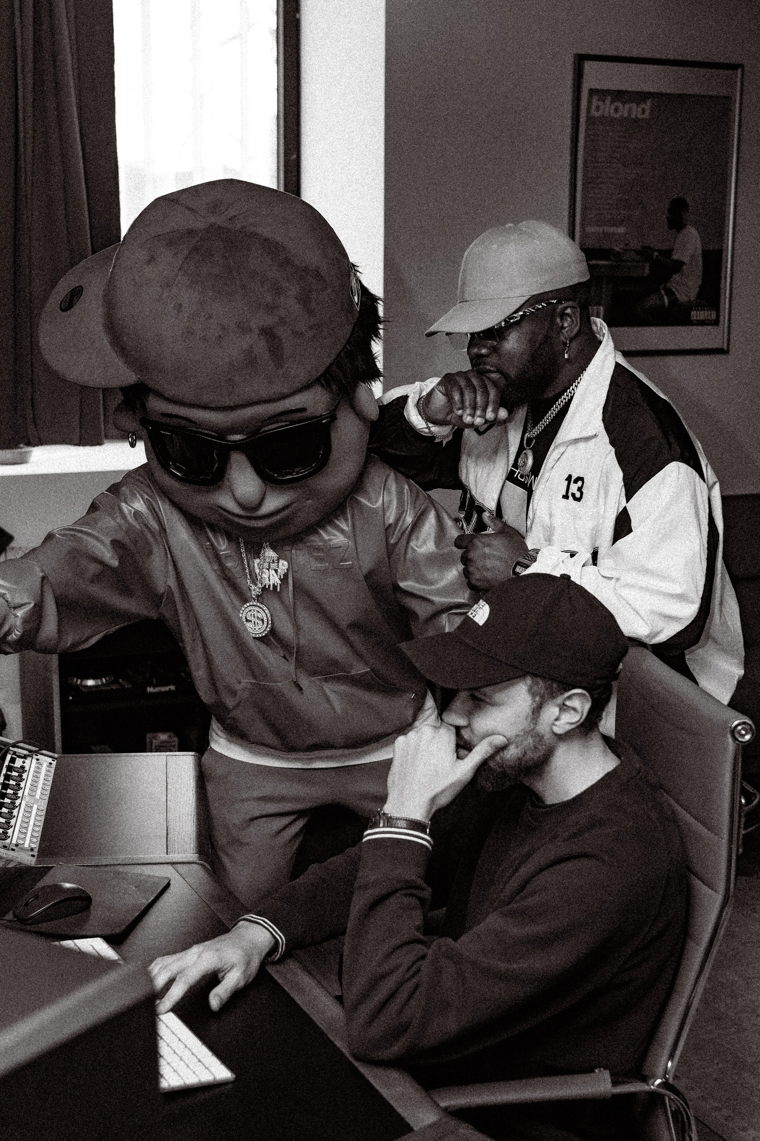 Trois hommes en studio d'enregistrement, deux debout et un assis, semblent être en pleine conversation ou interaction, portant des vêtements décontractés et des accessoires de mode, dans un style artistique noir et blanc.