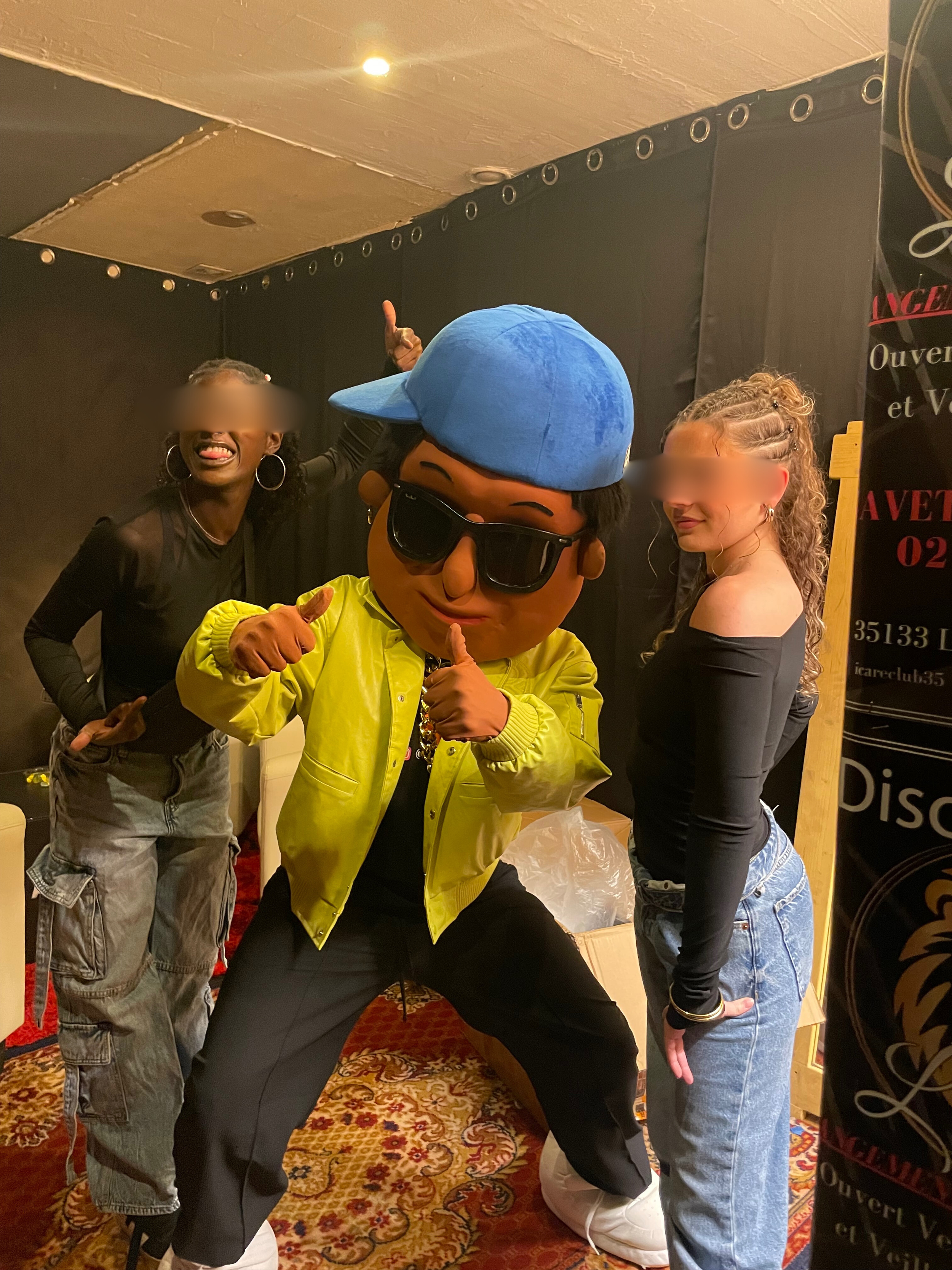 Scotti la mascotte avec un visage de personnage animé, portant une casquette bleue, des lunettes de soleil noires, une veste jaune, et faisant un signe de pouce en l'air accompagné de deux danseuses à la discothèque l'icare club