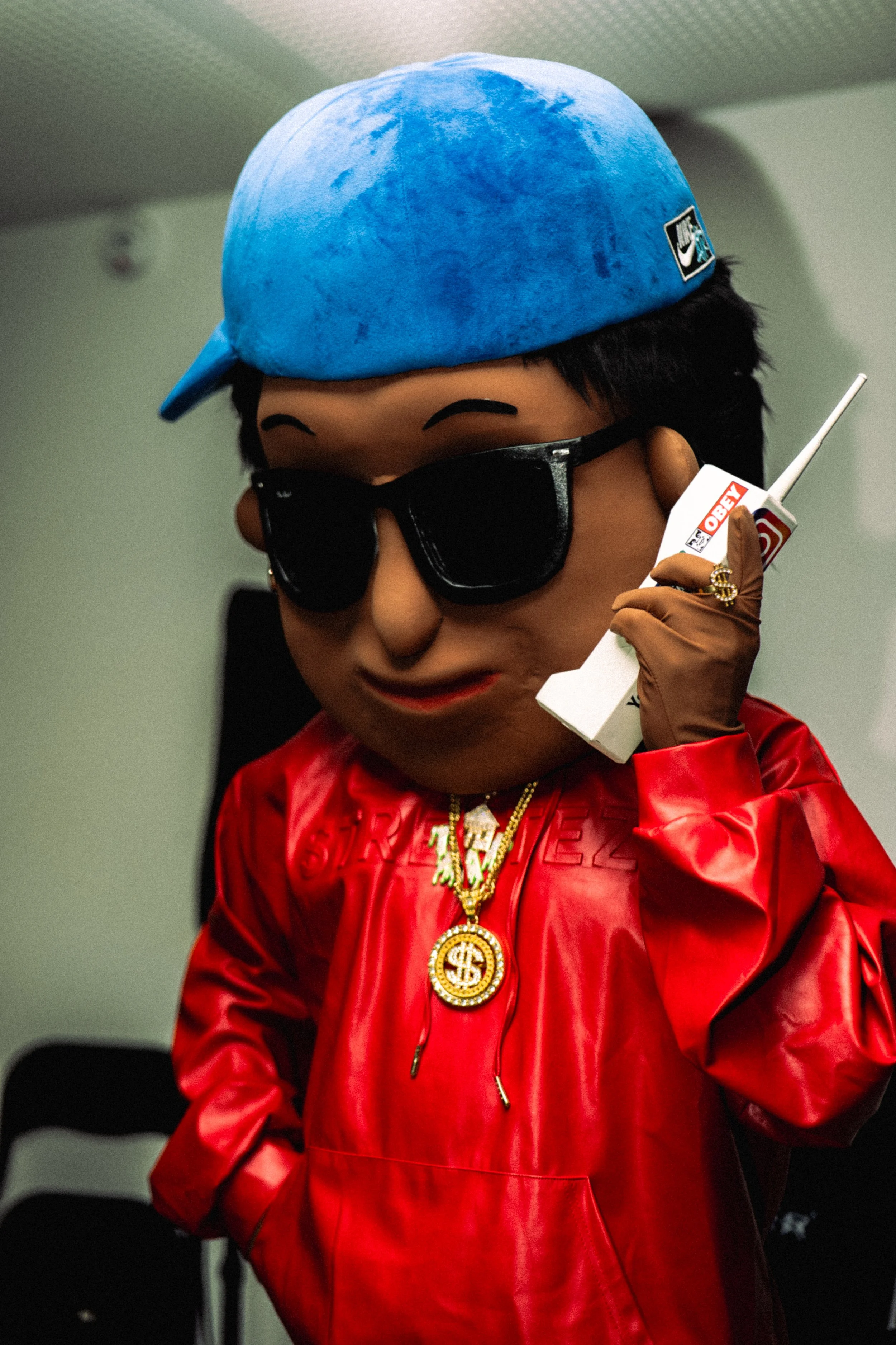 Mascotte SCOTTI portant une casquette bleue, des lunettes de soleil noires, une veste rouge en simili cuir et des bijoux dorés, tenant un téléphone en forme de boîte à cigarettes avec un logo "OBEY".