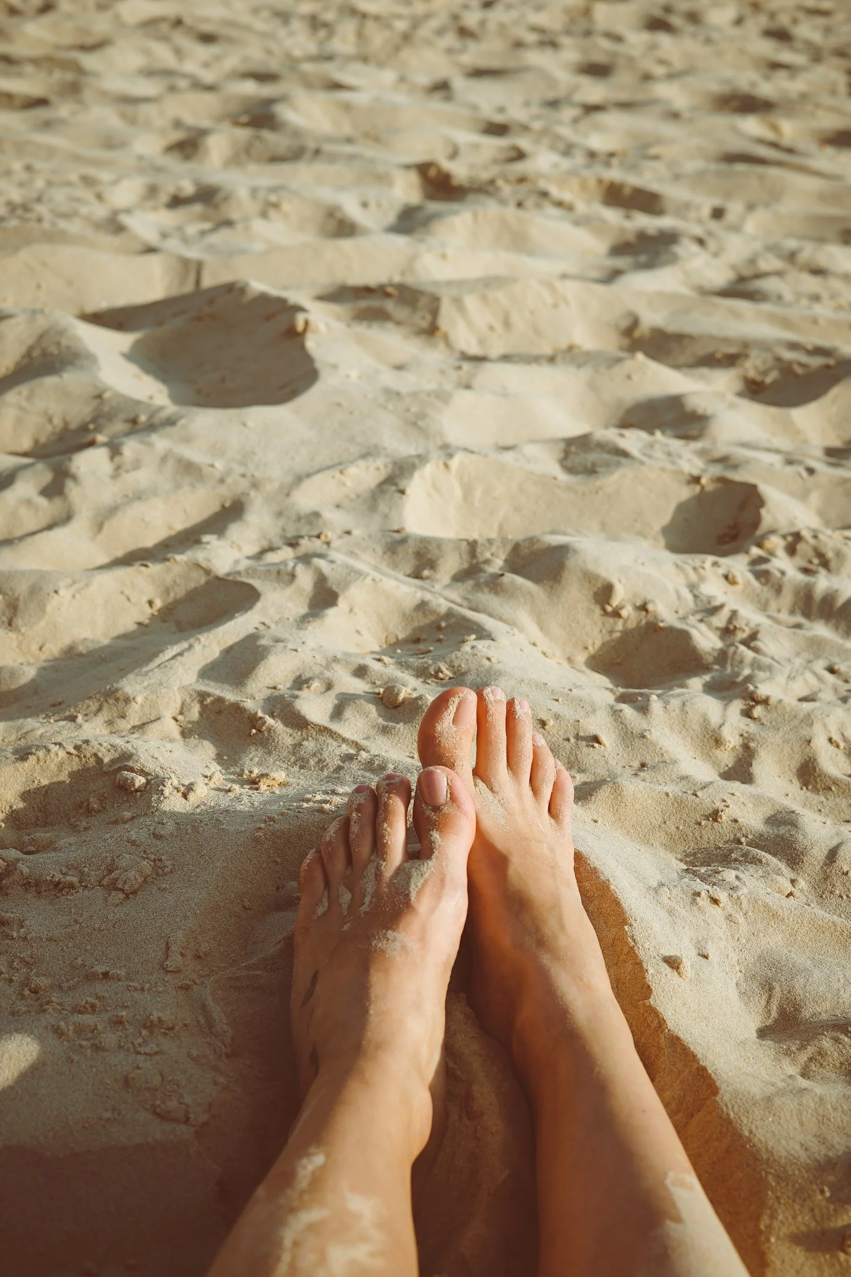 A pair of sandy feet on a sandy beach with footprints and sand dunes in the background.