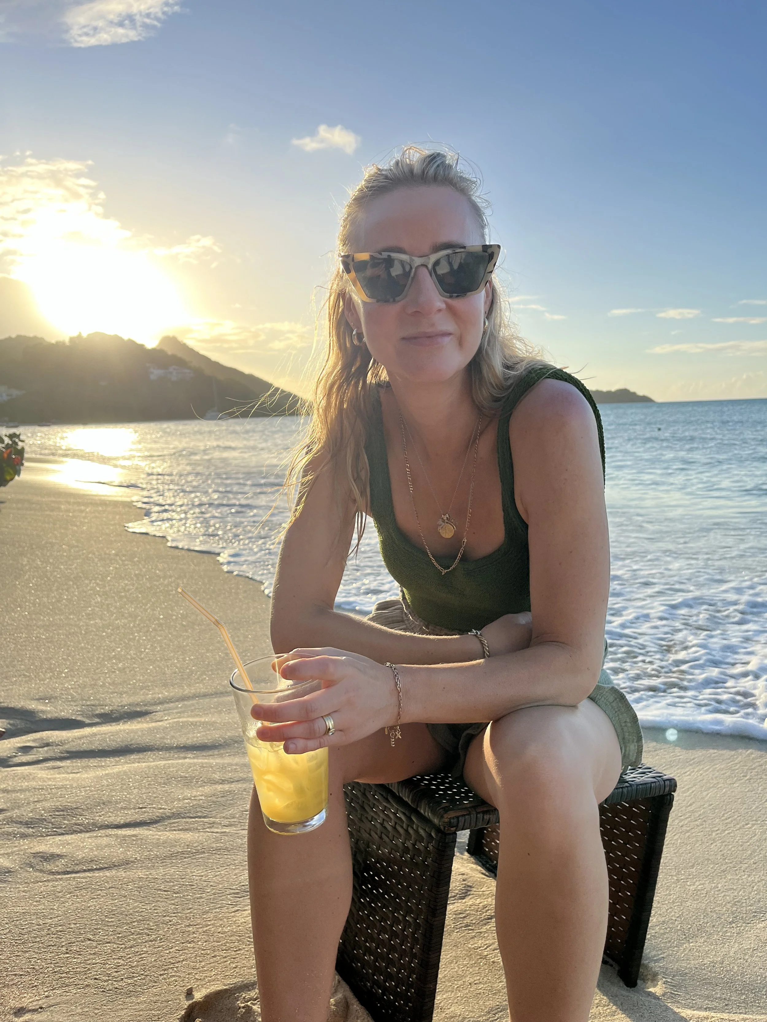 Woman sitting on a beach chair with a drink, wearing sunglasses, during sunset by the ocean.