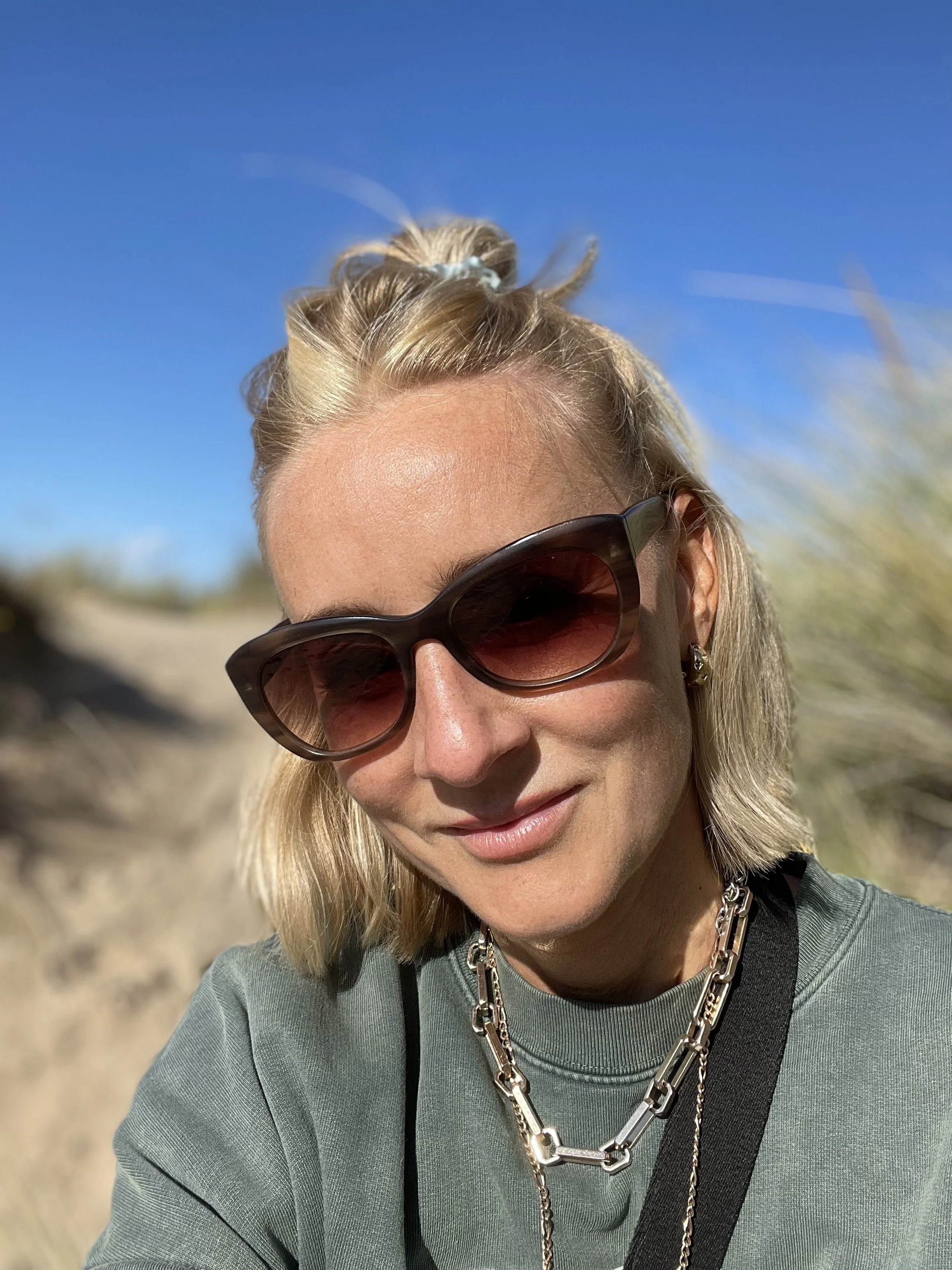 A woman with blonde hair, wearing large sunglasses, a green top, and layered gold jewelry, outdoors on a sunny day with a clear blue sky in the background.