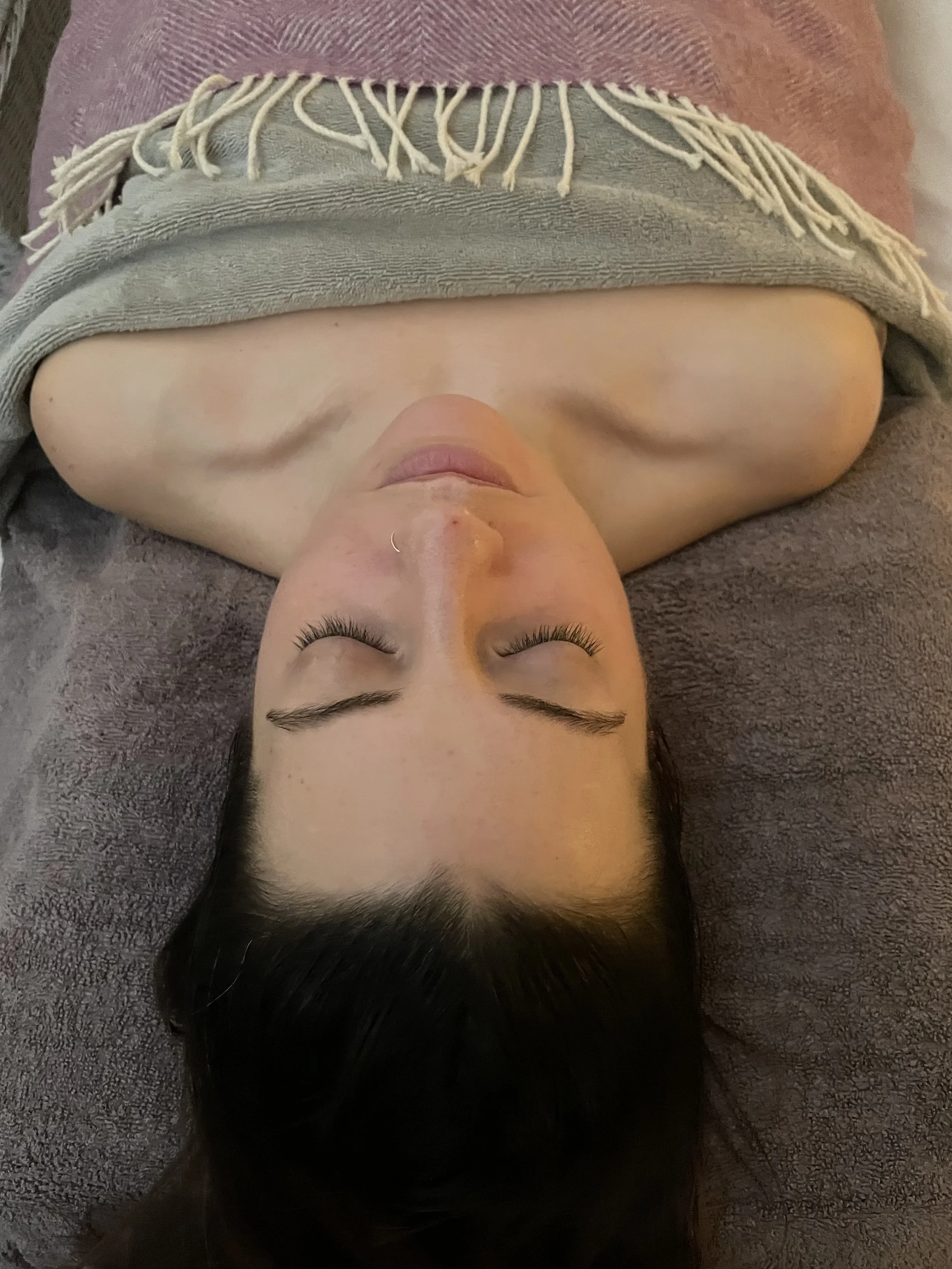 A woman lying on her back with her eyes closed, covered partially with a gray towel that has fringe at the ends, on a soft surface.