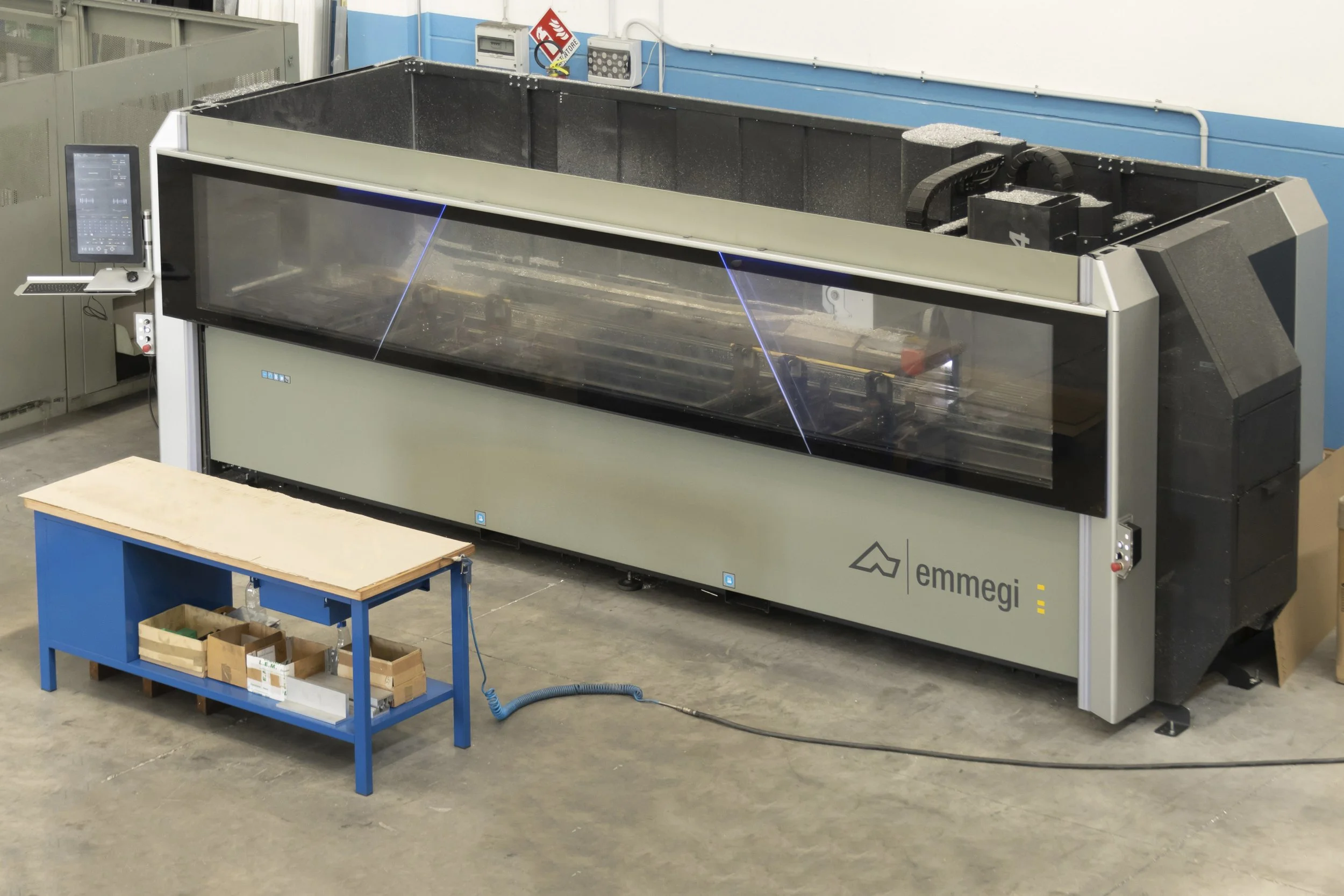 Una grande macchina industriale di colore beige e nero, con una superficie trasparente, situata in un ambiente di fabbrica con pavimento di cemento. Accanto c'è un tavolo blu con vari scatoloni e attrezzi.