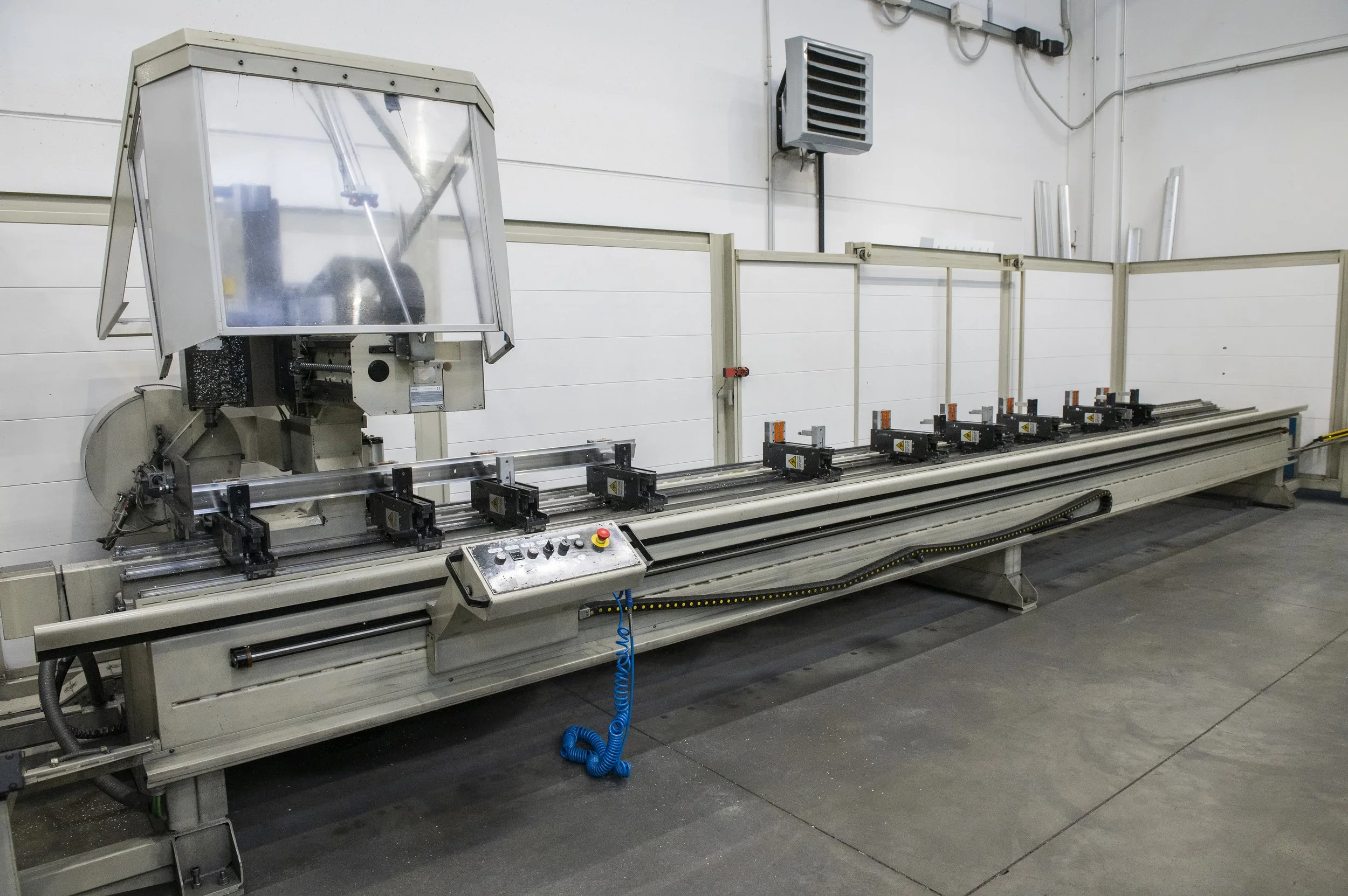Macchina industriale lunga con vari dispositivi di fissaggio, controlli e una copertura di protezione, situata in un ambiente industriale.