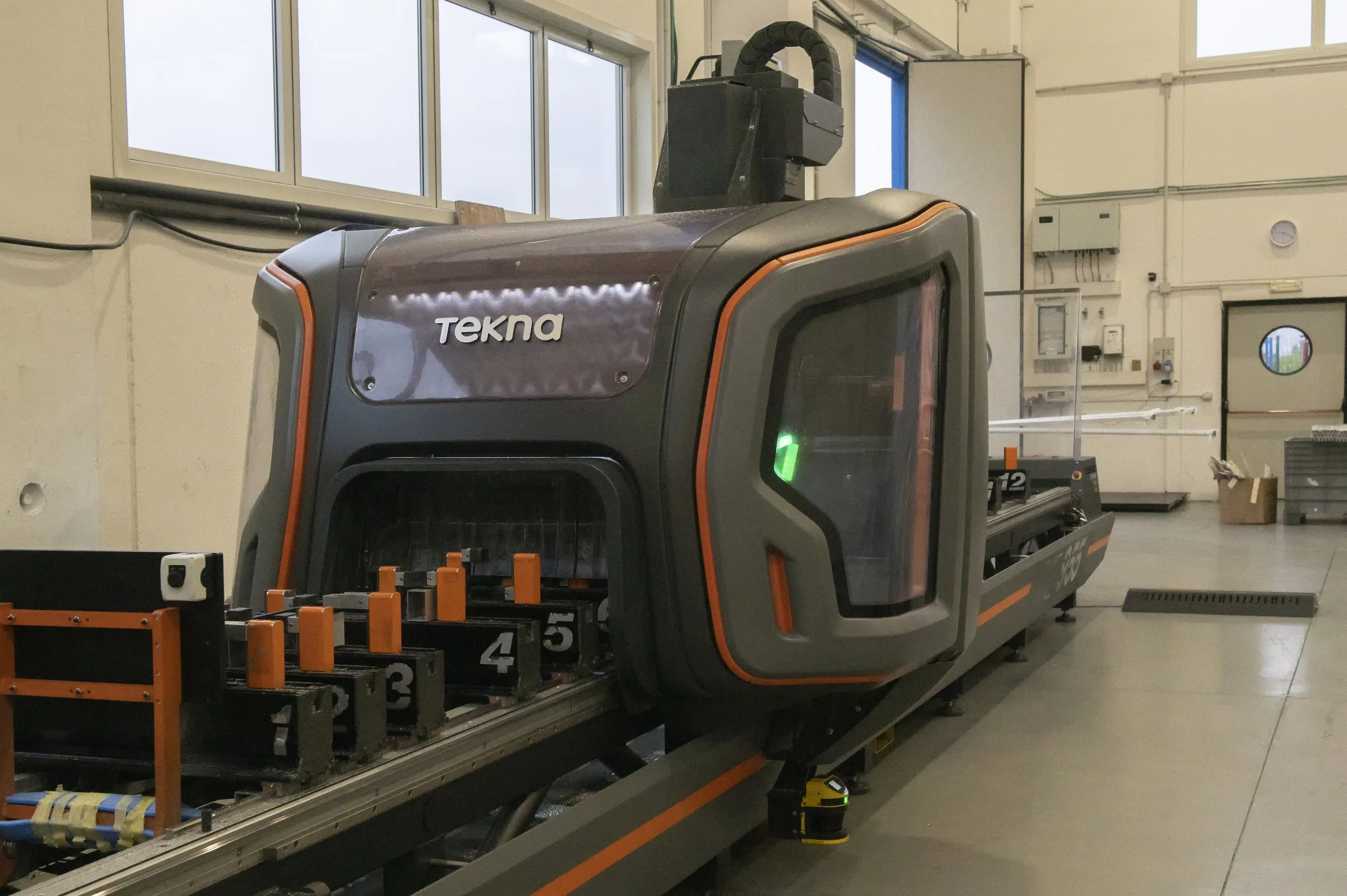 Un robot industriale di colore nero e grigio con dettagli arancioni, marca Tekna, installato in un ambiente di laboratorio o industriale, con finestre e pareti rivestite di attrezzature elettriche e strumenti.