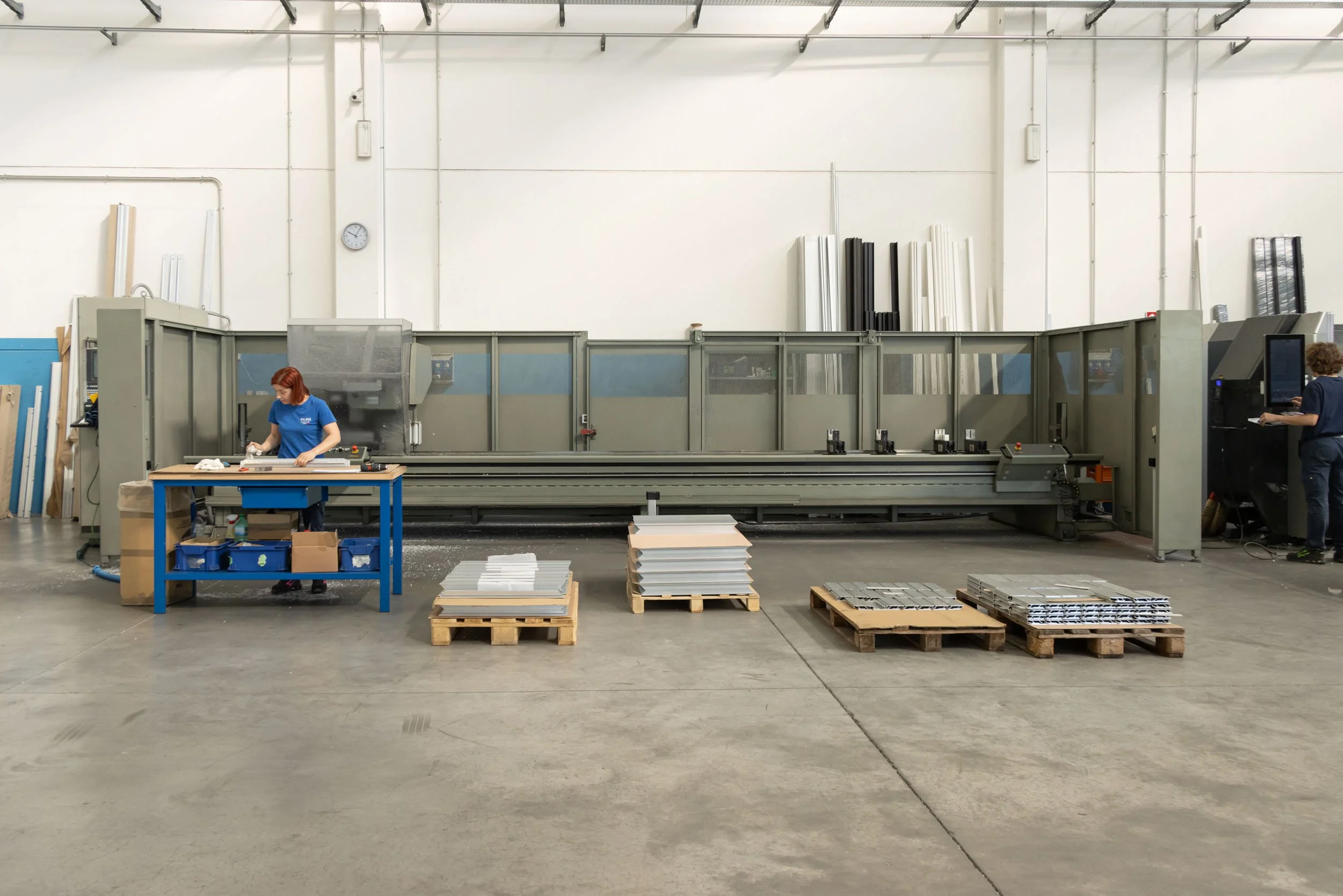 Interior di una fabbrica con grandi macchinari industriali, due persone che lavorano, e vari materiali e prodotti in lavorazione disposti su pallets di legno.
