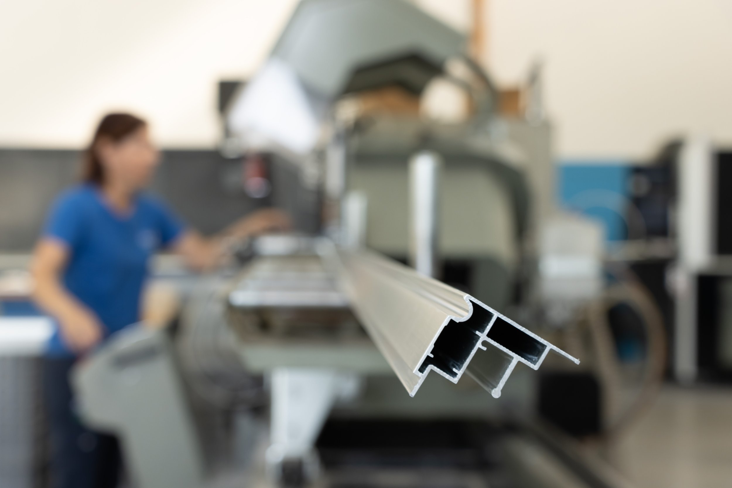 Dettaglio di un profilato di alluminio in primo piano, con una donna che lavora in un laboratorio industriale sfocata sullo sfondo.