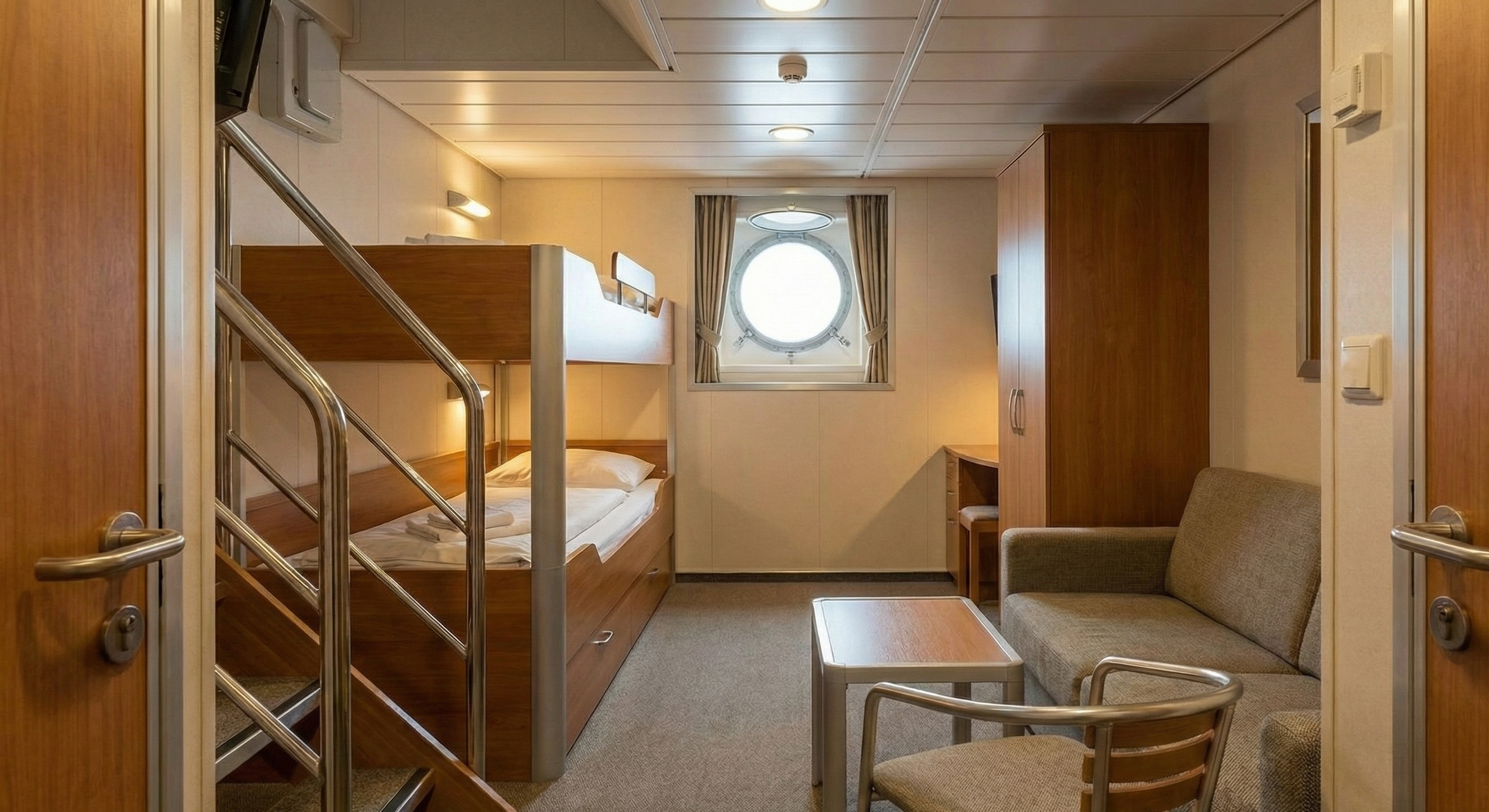 Interno di una cabina di nave con letti a castello, finestra rotonda, armadio, tavolino, due sedie e divano, in stile semplice e funzionale.