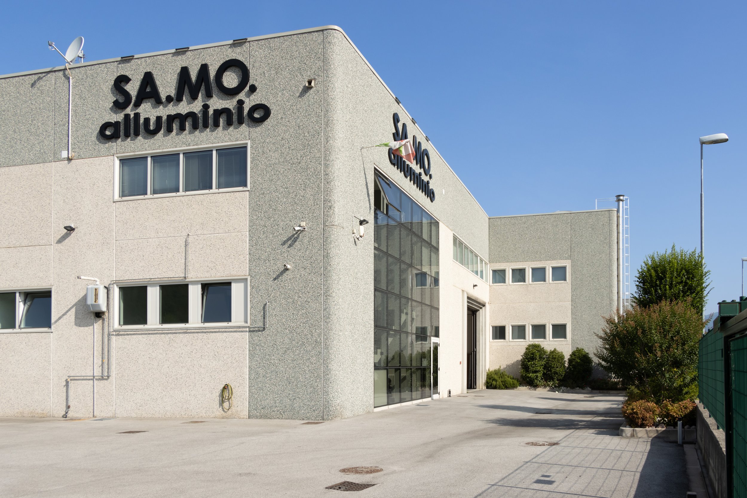Edificio industrial con le insegne 'S.A.M.O. alluminio' e pareti di rivestimento in gres, con alcune finestre e un cancello di ingresso, su un cielo blu e con vegetazione circostante.