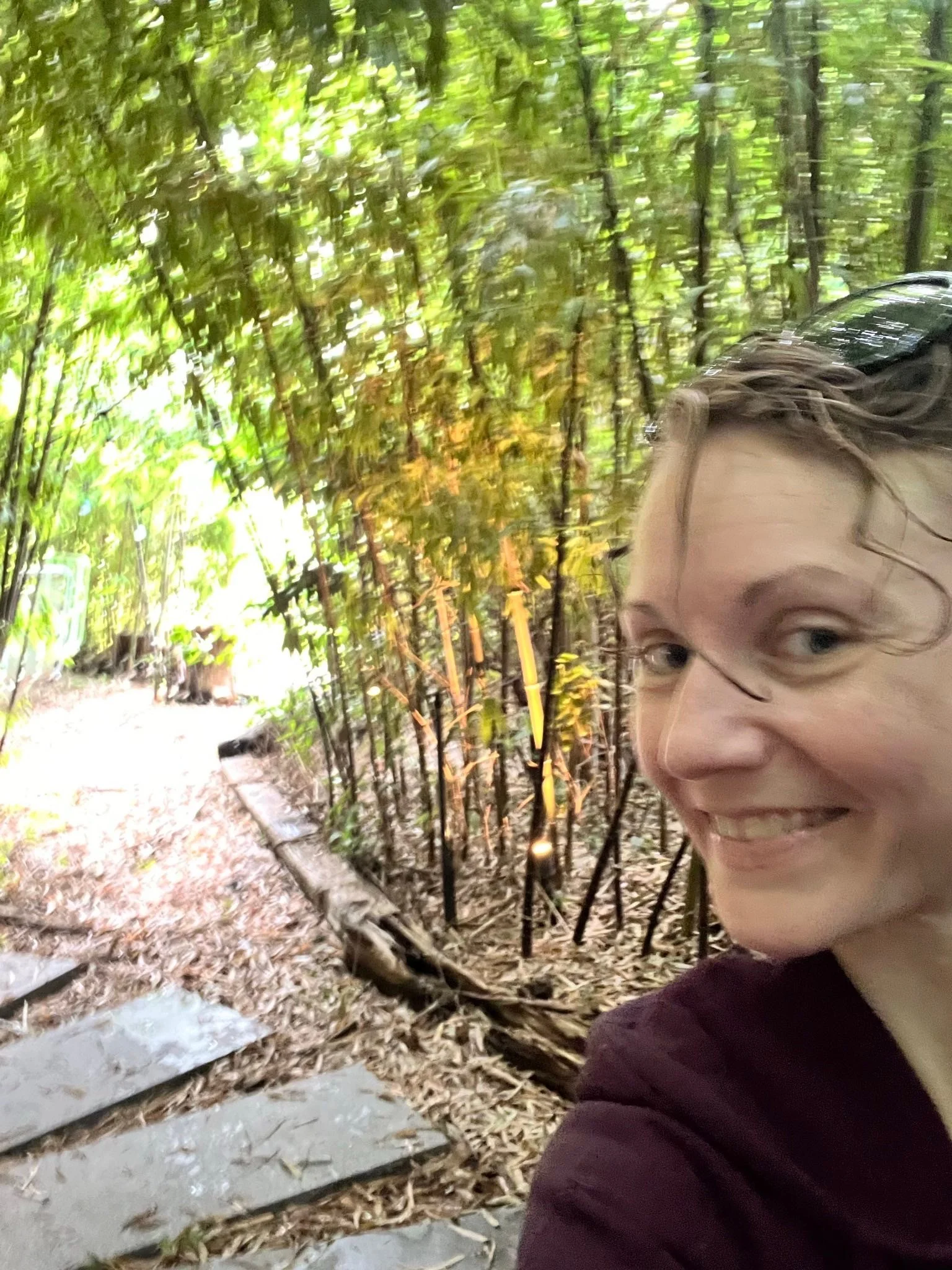 A grinning Anna leaning in for a selfie in front of rock stepping stone pathway through a bamboo forest.