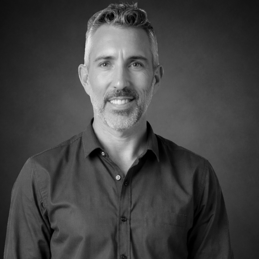 Black and white professional portrait of a middle-aged man with gray hair and beard, wearing a dark button-up shirt, smiling softly against a plain background.