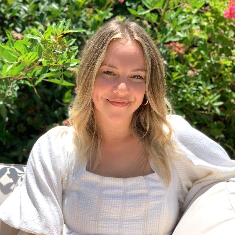 A woman with blonde hair smiling outdoors, surrounded by green foliage and pink flowers, wearing a white top.