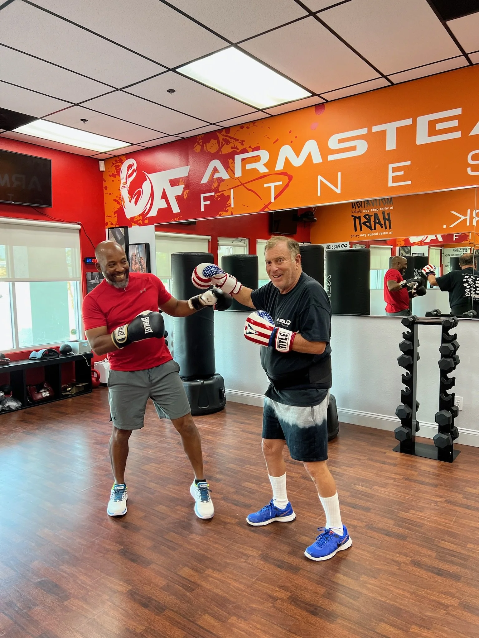 Two men in a gym, smiling and boxing with gloves, standing on a wooden floor near punching bags, with a mirror reflecting their image and a large orange and red sign reading 'Armstead Fitness' on the wall.