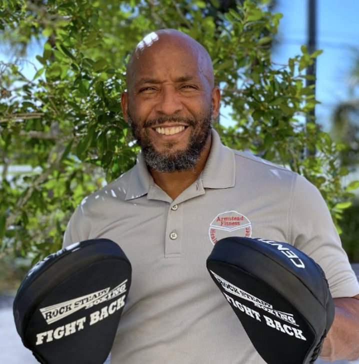A smiling man with a bald head and beard, wearing a gray polo shirt with 'Armstead Fitness' logo, holds boxing gloves that say 'Rock Steady Boxing Fight Back,' standing outdoors with green foliage in the background.