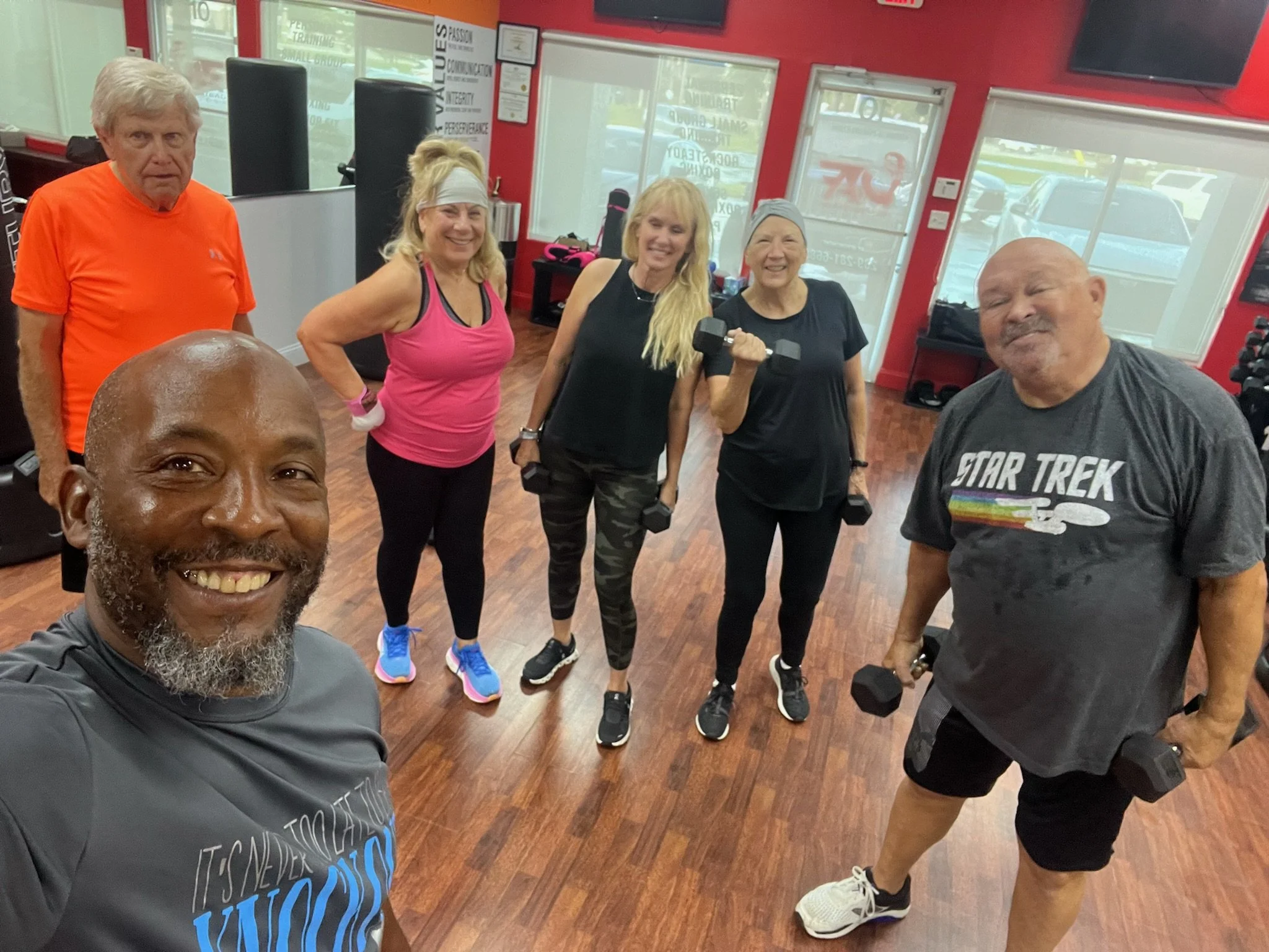 Group of six older adults in a gym, smiling for a selfie, holding dumbbells, with a red wall and large windows in the background.