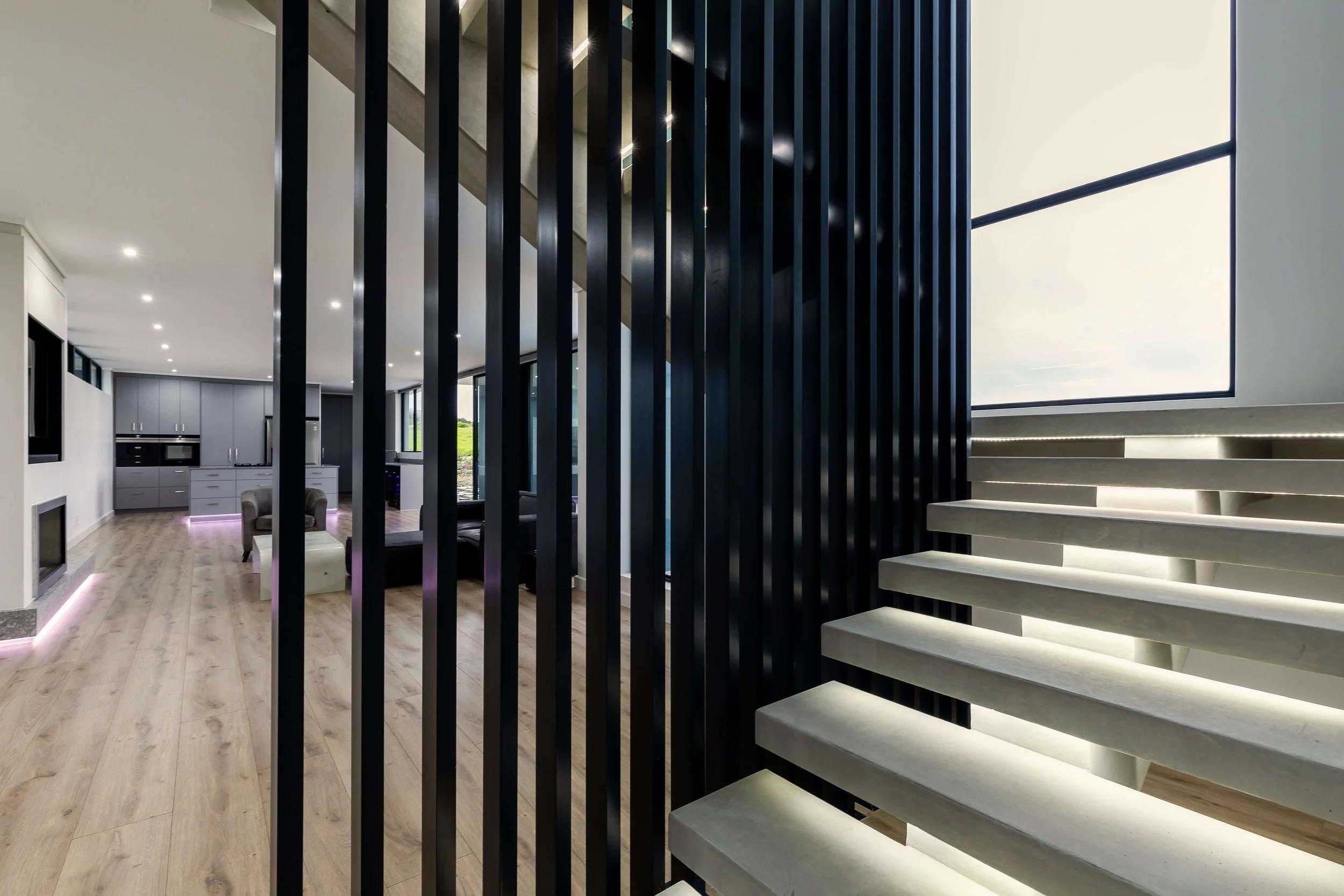 Modern interior with a staircase featuring illuminated light strips, black vertical slats, and a view of the open-plan living area with kitchen and seating.