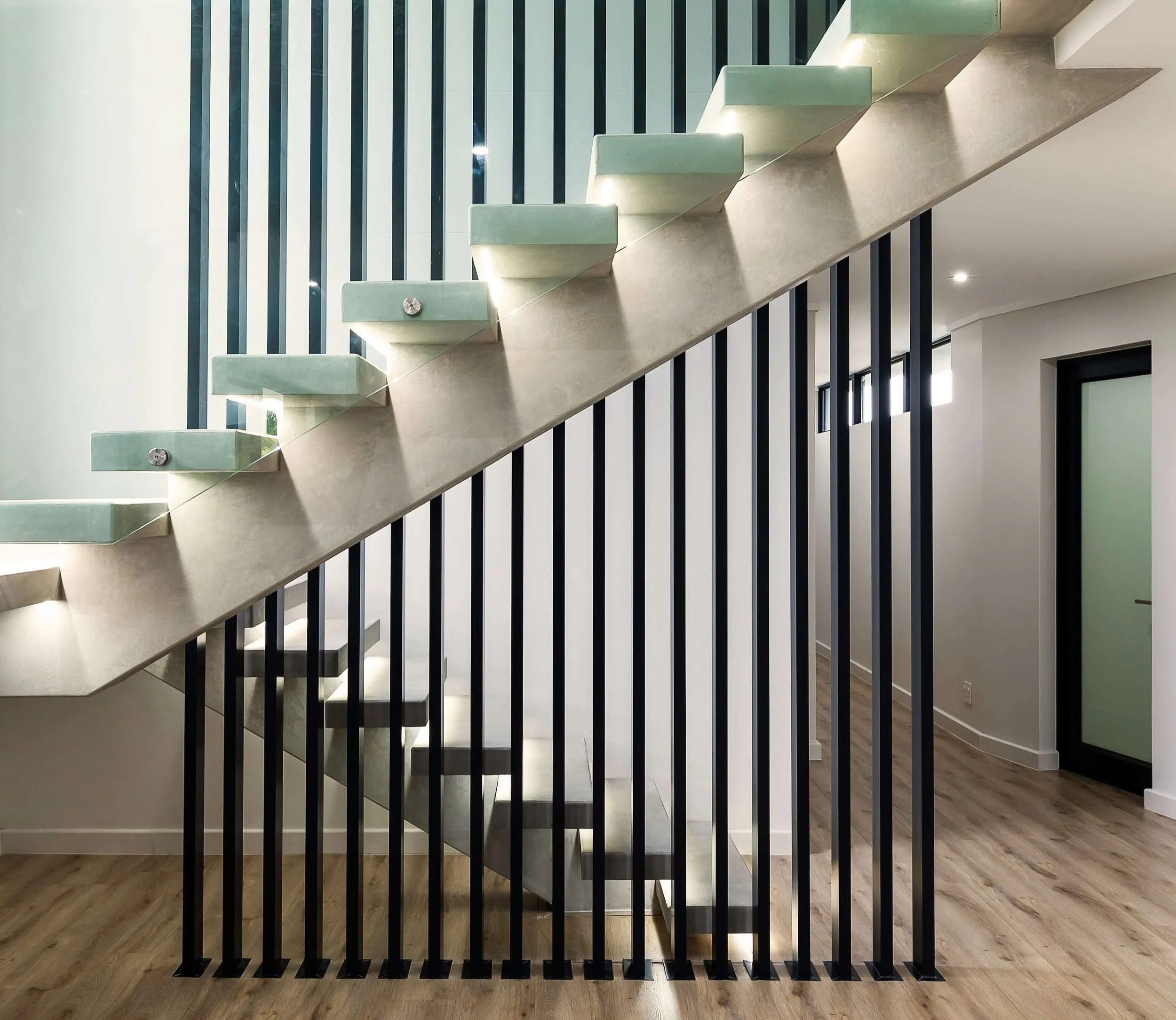 Modern interior staircase with floating light-colored concrete steps and black vertical metal balustrades, in a spacious, well-lit room with light wood flooring and white walls.