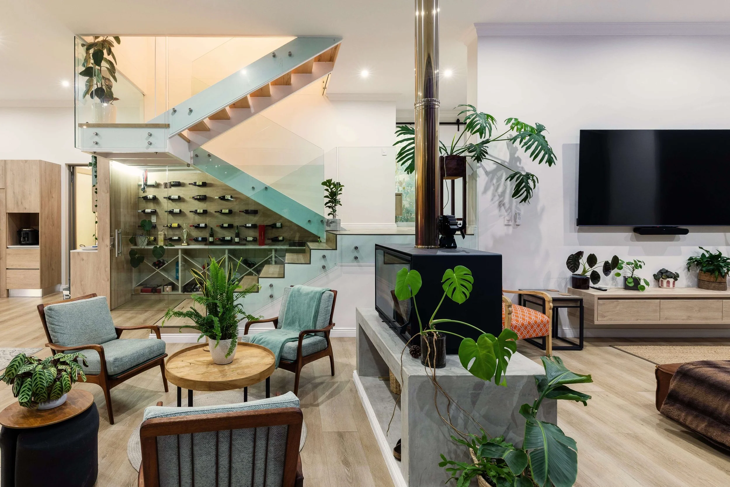 Living room interior with modern decor, wooden furniture, green plants, a large flat-screen TV on the white wall, and a staircase with glass railing leading to an upper level. There is a wine rack under the staircase.