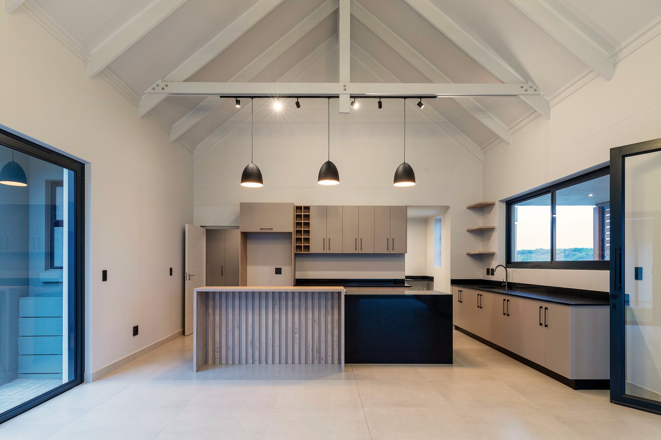 Modern kitchen with beige cabinets, black countertops, and large windows, featuring a central island with a striped wooden front, black pendant lights, and open shelving on the wall.