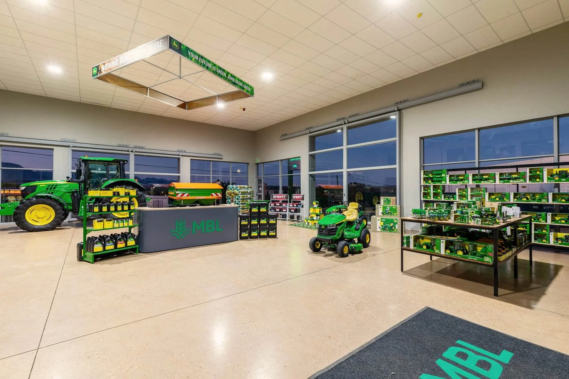 Inside a John Deere store displaying green and yellow lawn equipment and accessories, including a large tractor, a riding mower, and shelves stocked with toy models of John Deere equipment.