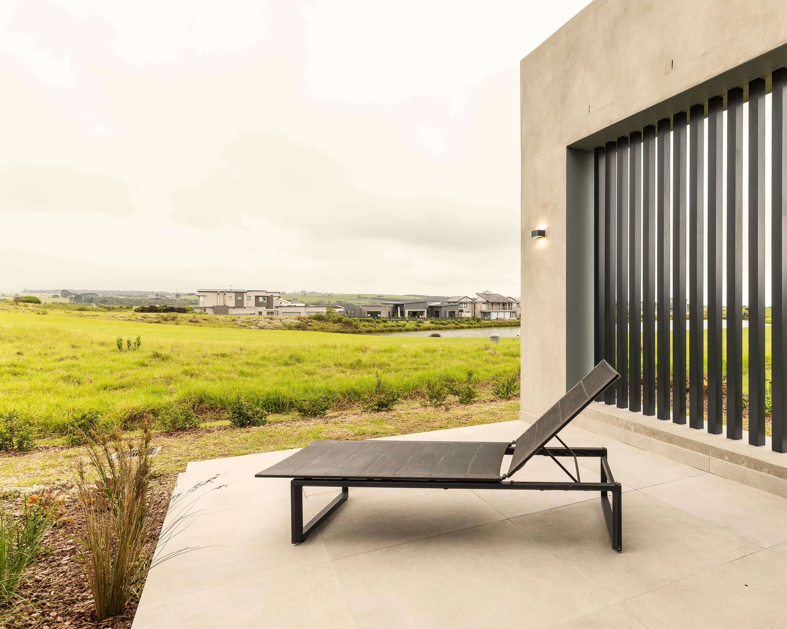 An outdoor patio with a lounge chair overlooking a grassy field and modern houses in the distance.
