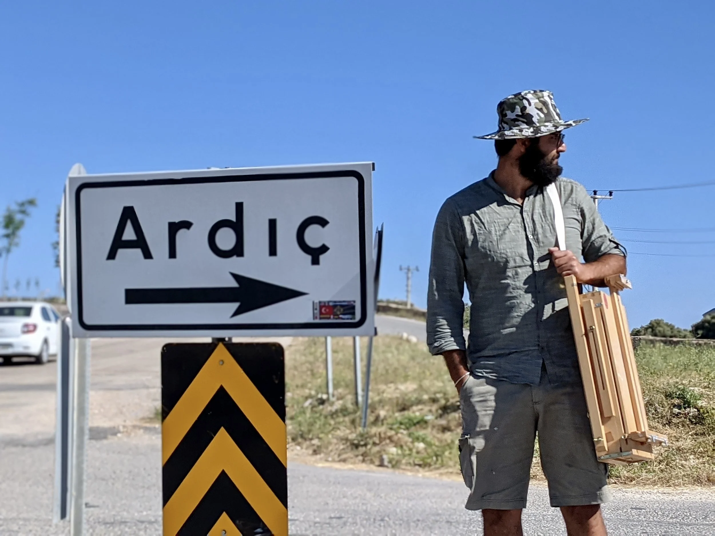 Man with a beard wearing a camouflage hat, sunglasses, gray shirt, and shorts, holding a wooden item, standing next to a road sign with the word 'Ardıç' and an arrow pointing right under a blue sky.