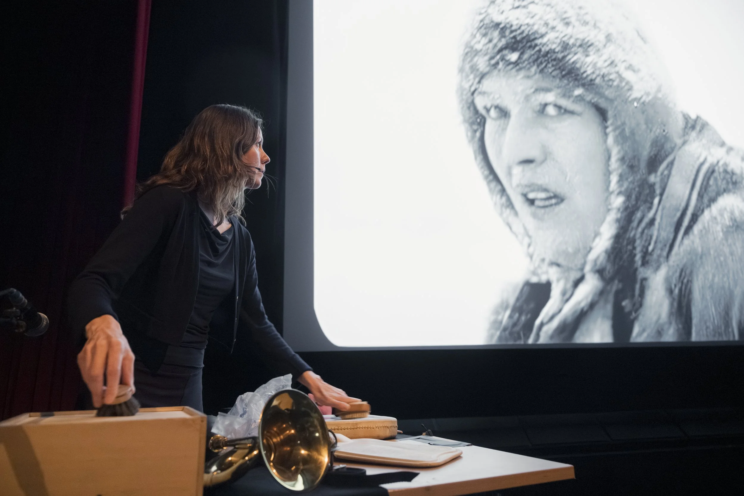 En kvinne står ved et bord med ulike objekter foran en stor skjerm som viser et svart-hvitt bilde av en person i vinterklær. Kvinnen vender mot skjermen og ser ut til å ta notater eller forberede noe.