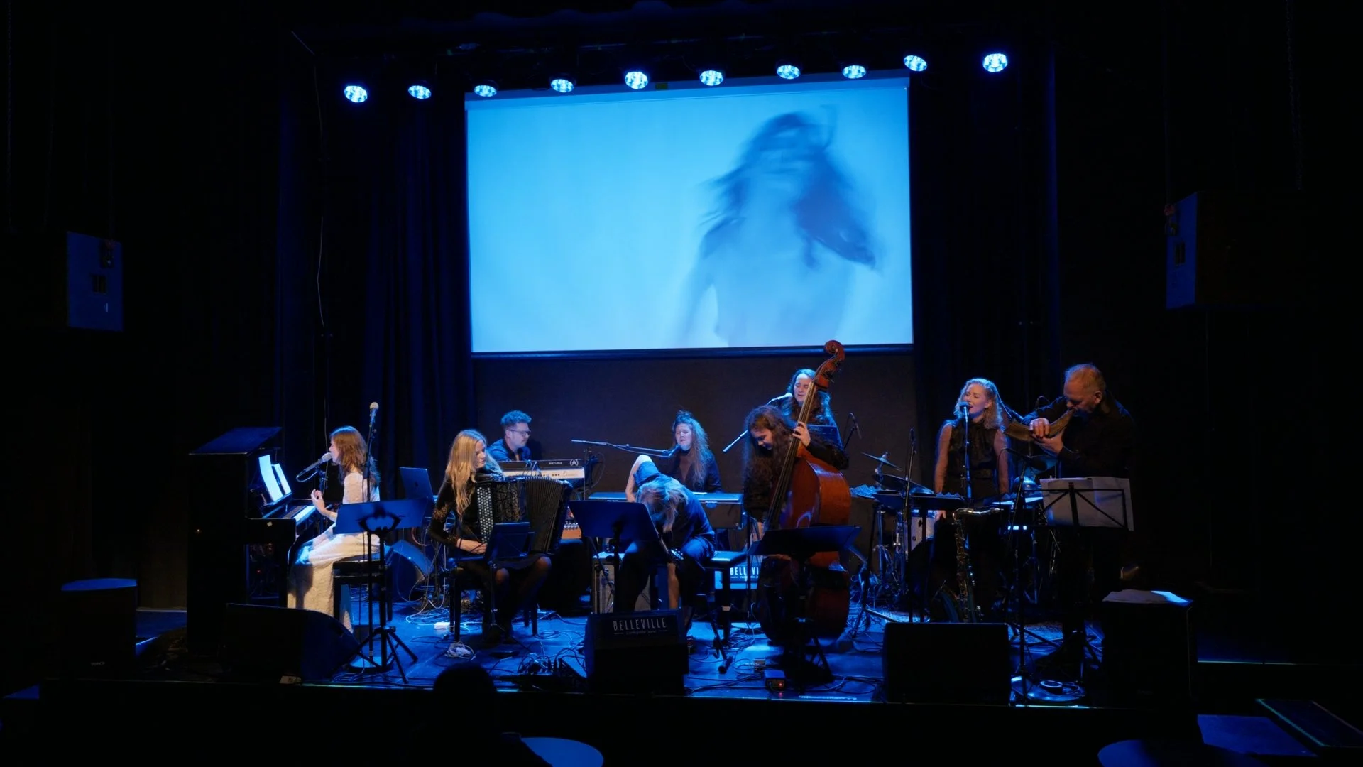 En musikalsk gruppe som spiller på scenen under en konsert, med et stort lerret bak dem som viser en vage, blåaktig stumfilm av en person. Gruppemedlemmene bruker ulike instrumenter som piano, accordion, kontrabass og fiolin.
