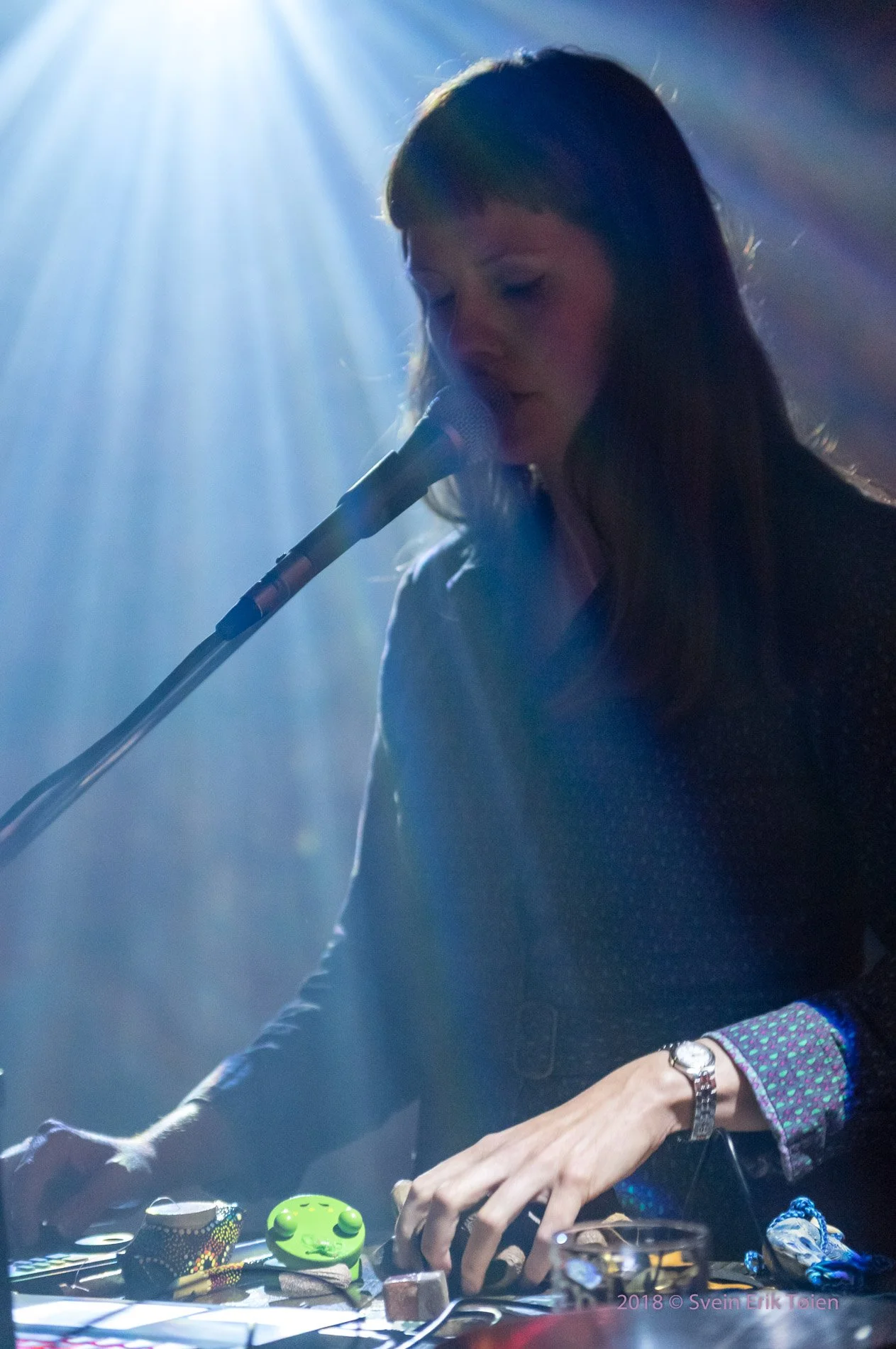 En kvinne som spiller elektroniske instrumenter på en scene med blå belysning og en mikrofon foran seg.