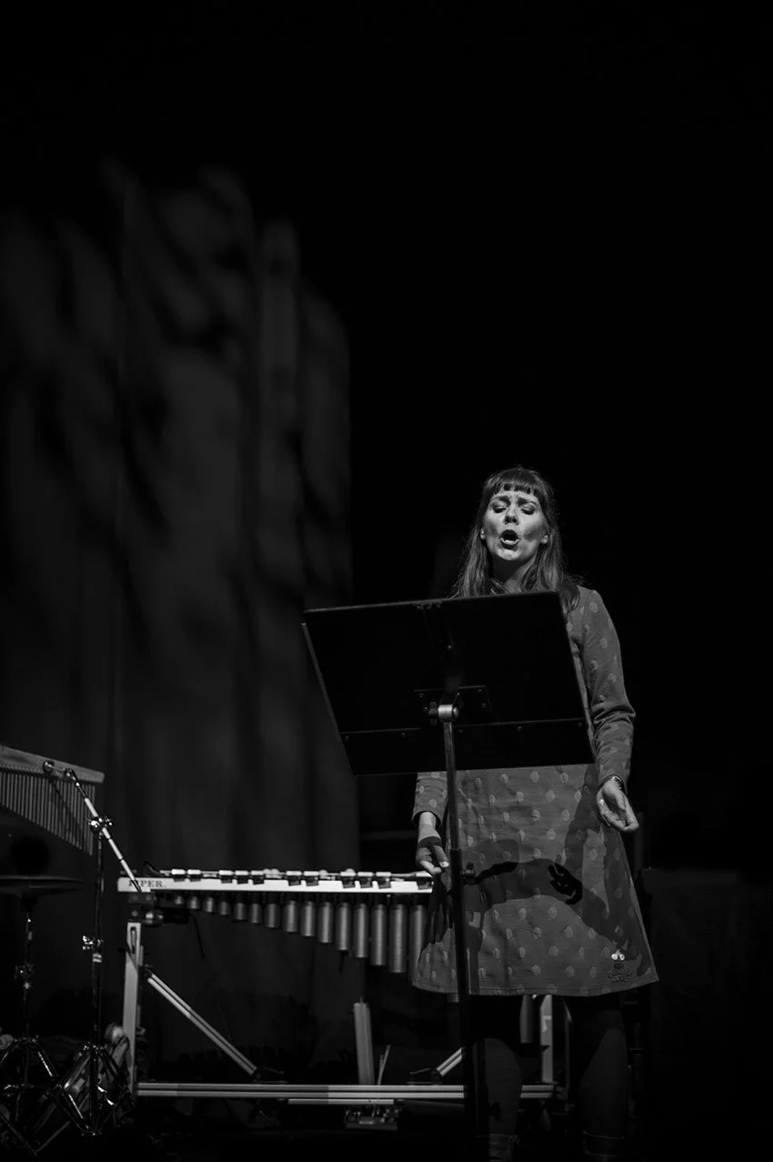 En kvinnelig sanger står foran en mikrofon på en scene, med en musikkstativ og et metallrør i forgrunnen. Det er svart bakgrunn.