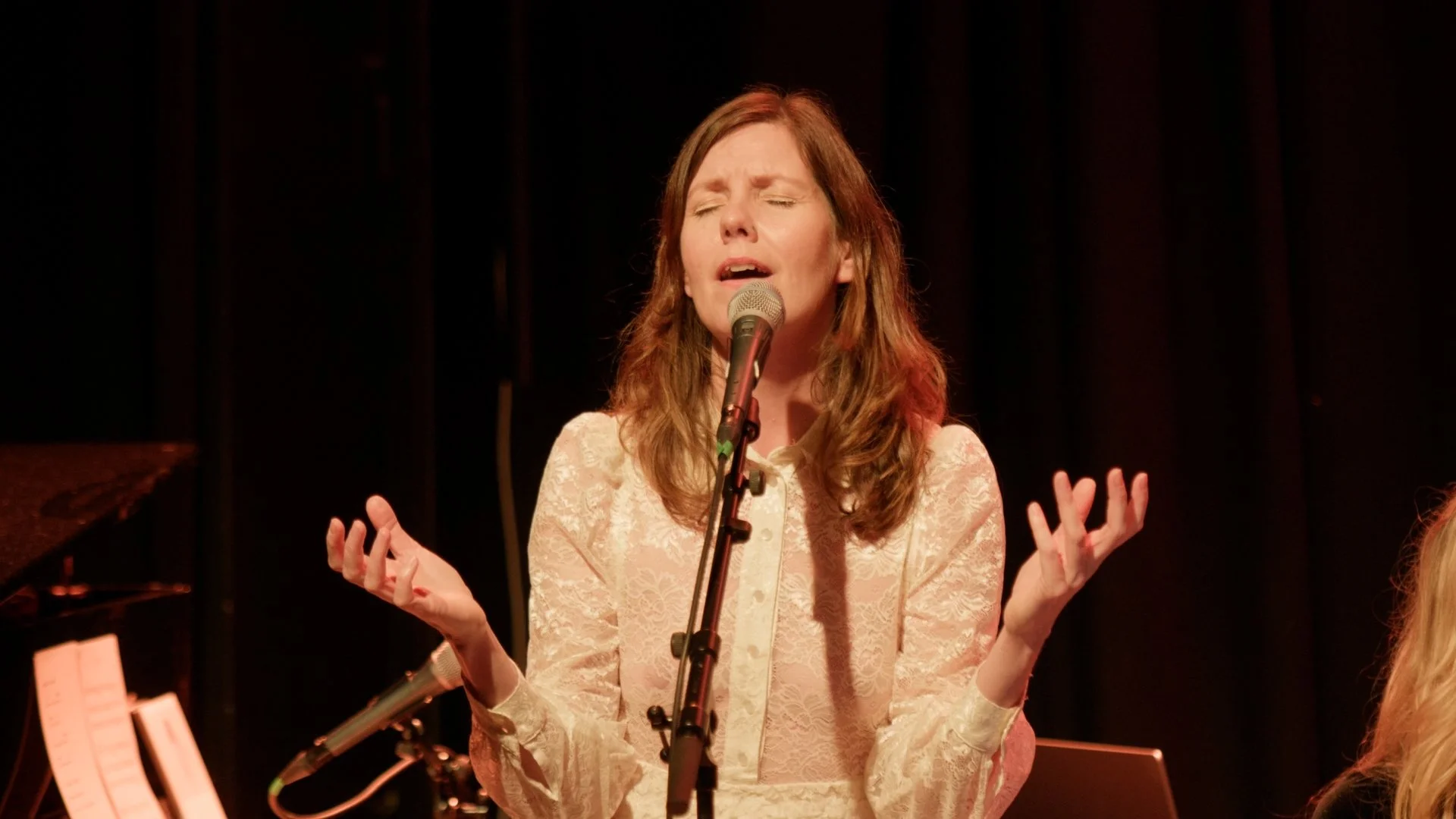 En kvinne synger med lukkede øyne, kledd i en elegant, lys blondedrakter, på scenen med en mikrofon, omgitt av musikkutstyr.