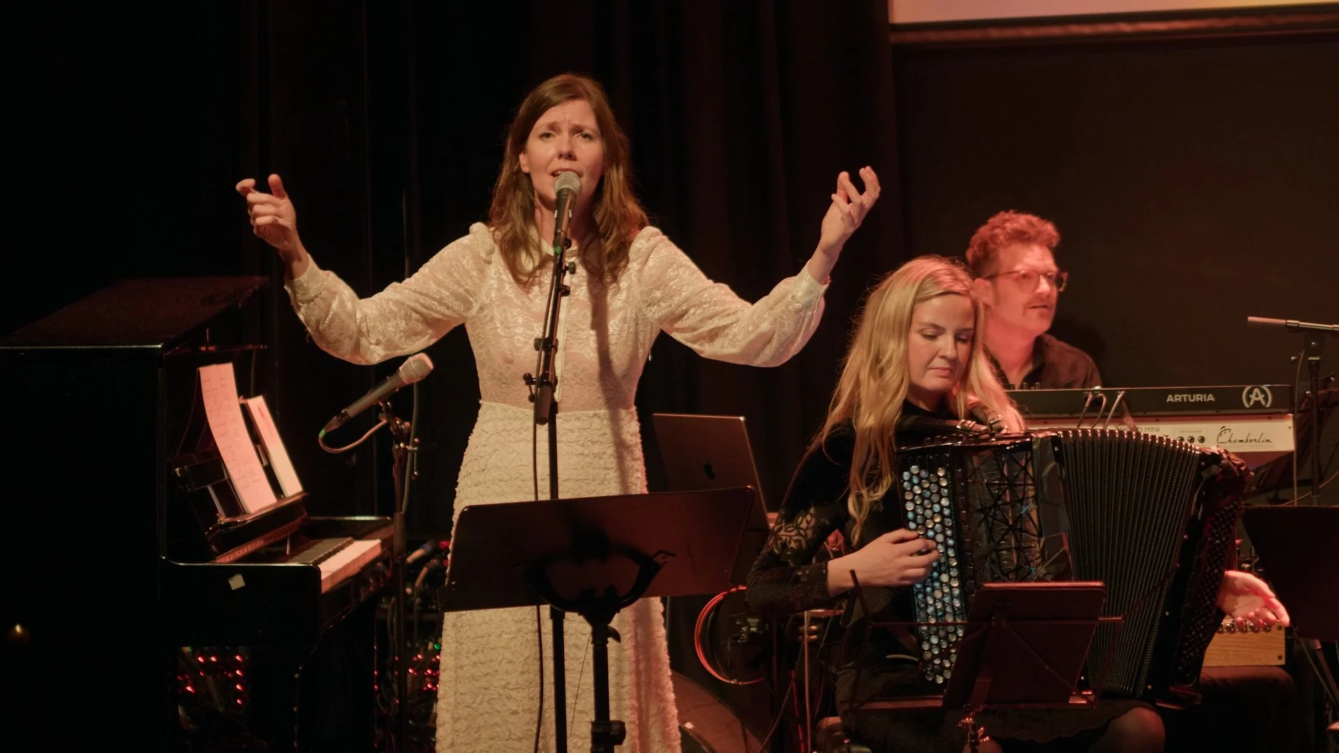 Tre musikere på scenen, en kvinne synger i mikrofon med armer utstrakt, to andre spiller instrumenter, en piano og en trekkspill, omgitt av musikkliver