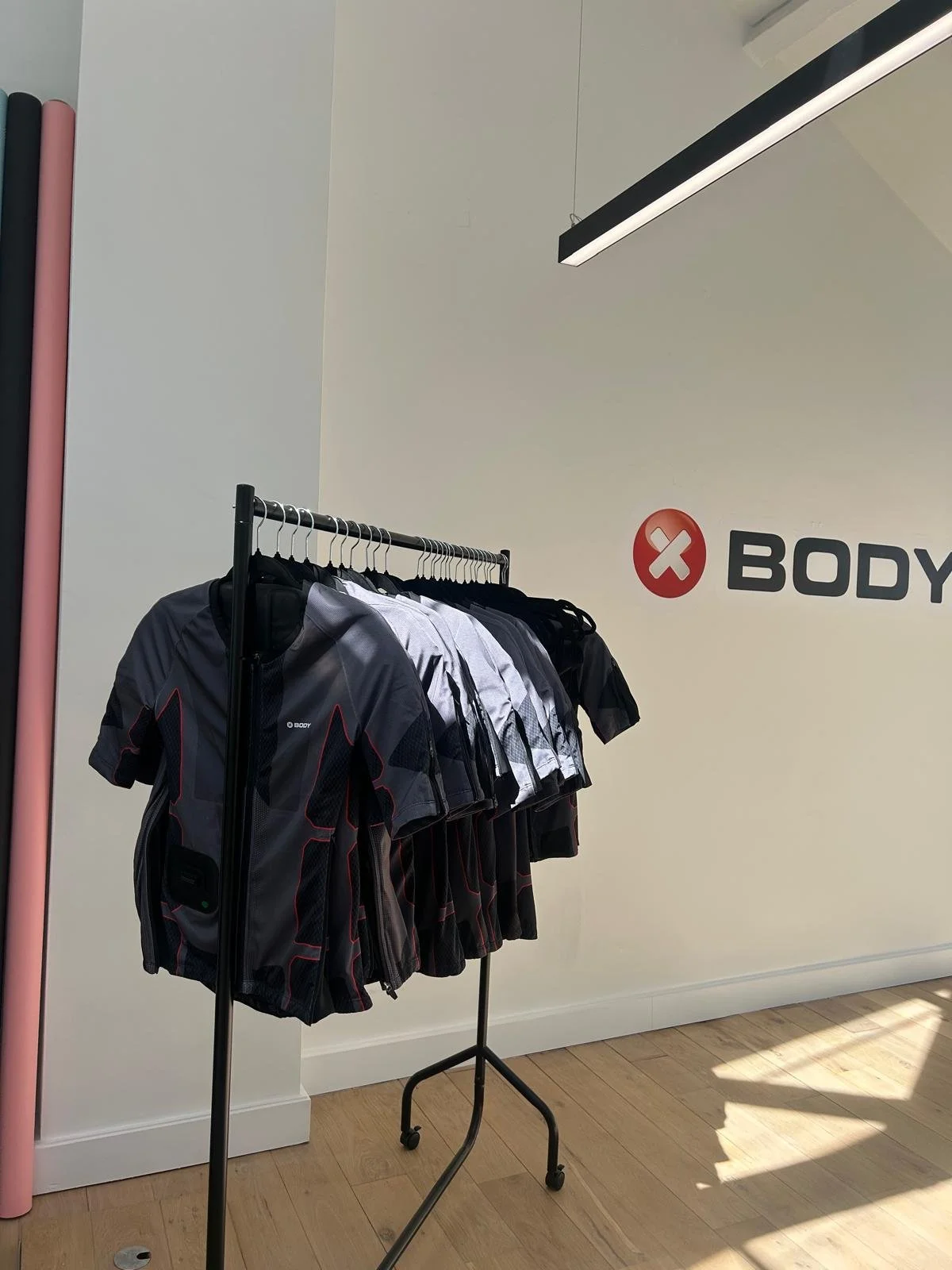 Clothing rack with athletic shirts in black, gray, and white, in a retail store with a white wall and wood floor, part of the word 'BODY' visible on the wall.