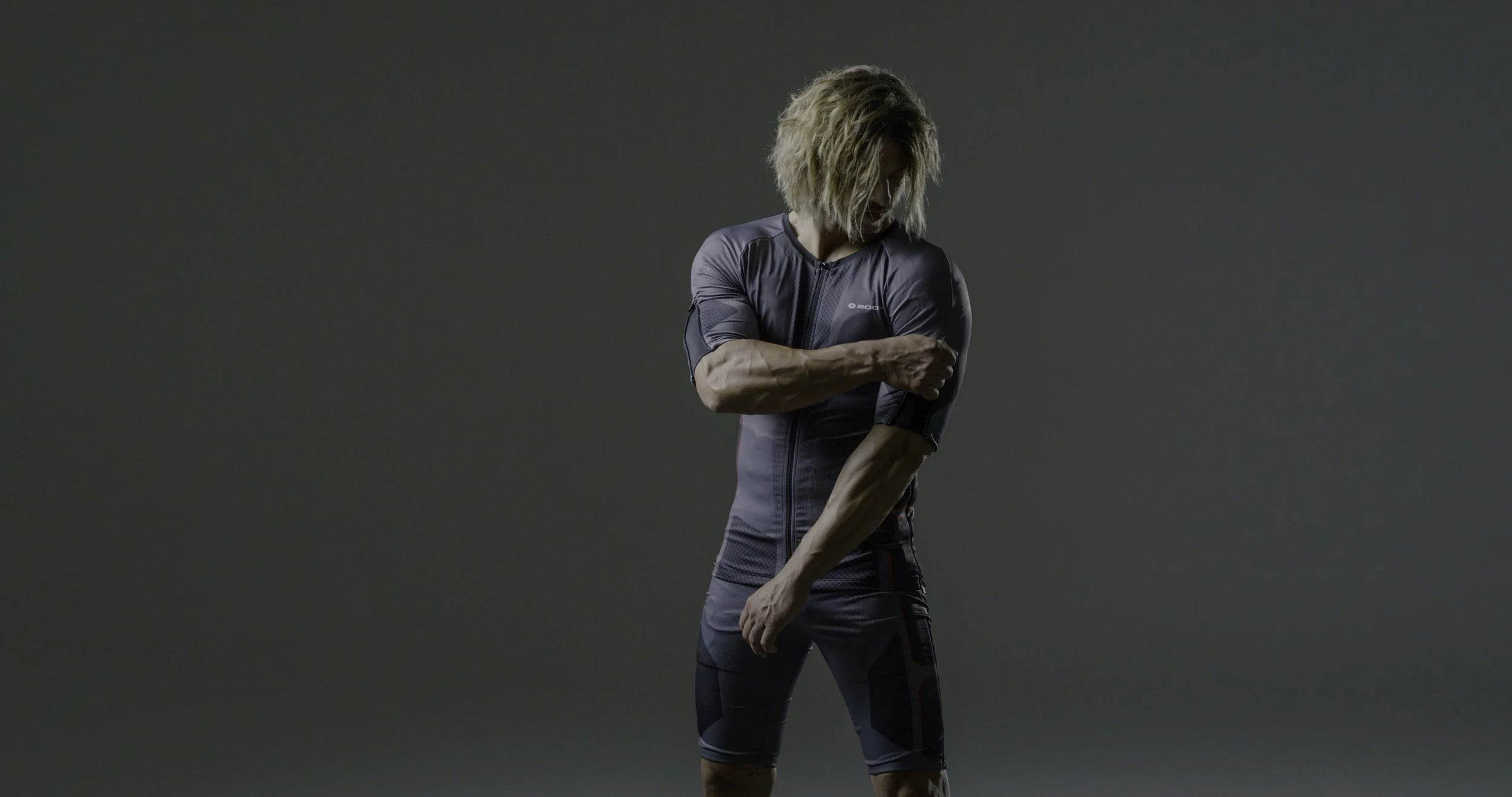 A person with curly blonde hair in athletic gear adjusting their sleeves against a plain gray background.