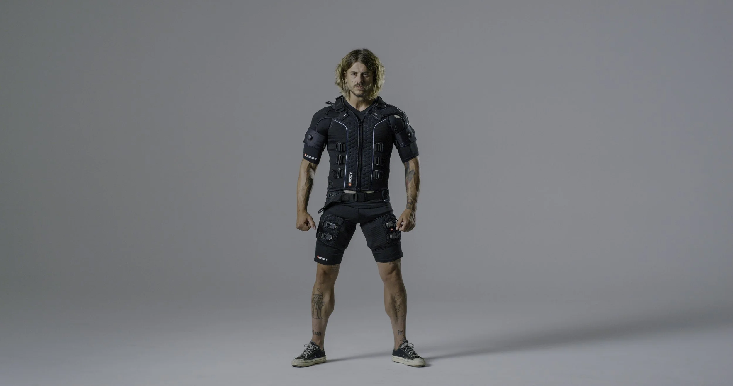 Man standing against a plain gray background wearing black tactical gear, shorts with multiple pouches, and sneakers, looking directly at the camera.