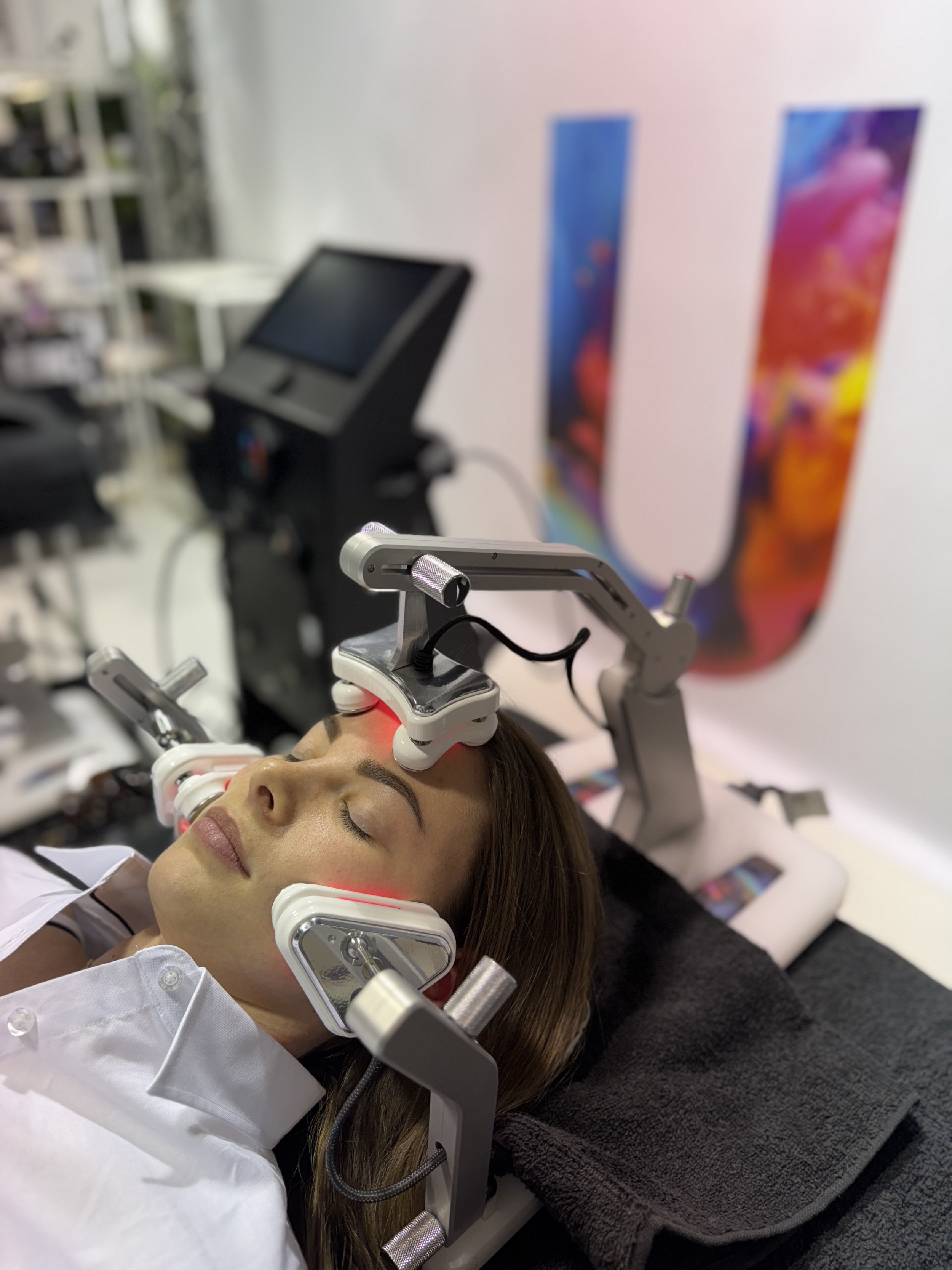 Woman lying down with her eyes closed during a cosmetic procedure using a laser device on her face, with a monitor and promotional posters in the background.