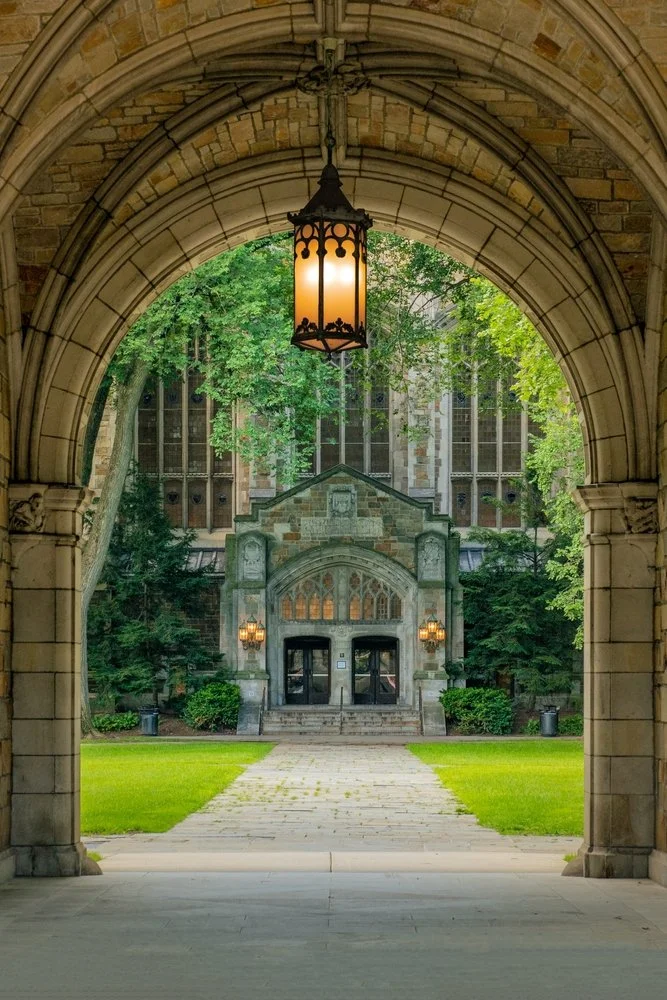 A stone archway with a hanging lantern, leading to a pathway through a grassy area, with trees and an older building with large windows in the background.