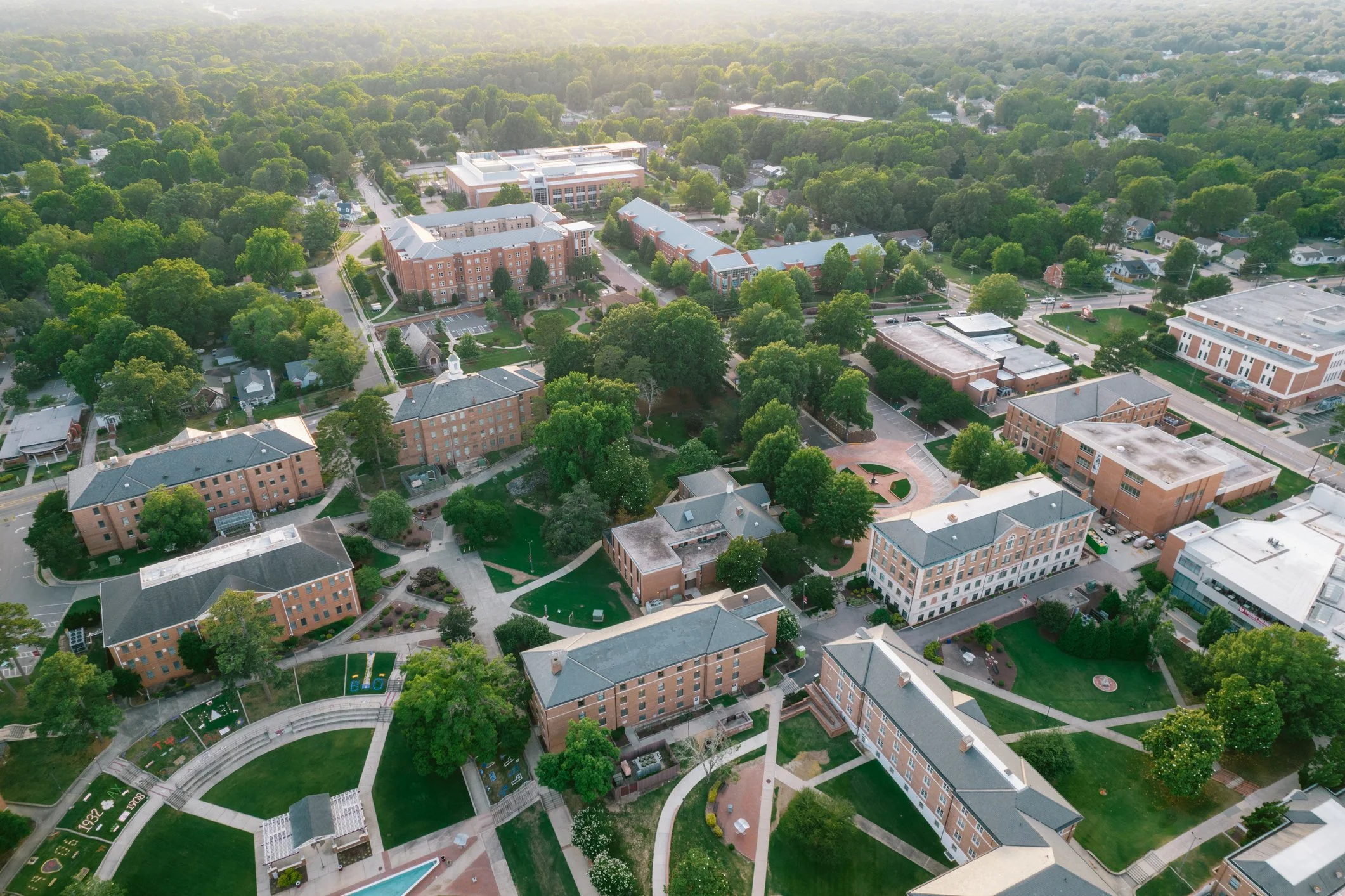 Aerial view of a college campus with brick buildings, green trees, pathways, and parks, surrounded by suburban houses and streets.