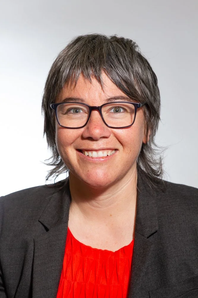 A woman with short gray hair, wearing glasses, a black blazer, and a red blouse, smiling at the camera against a plain gray background.
